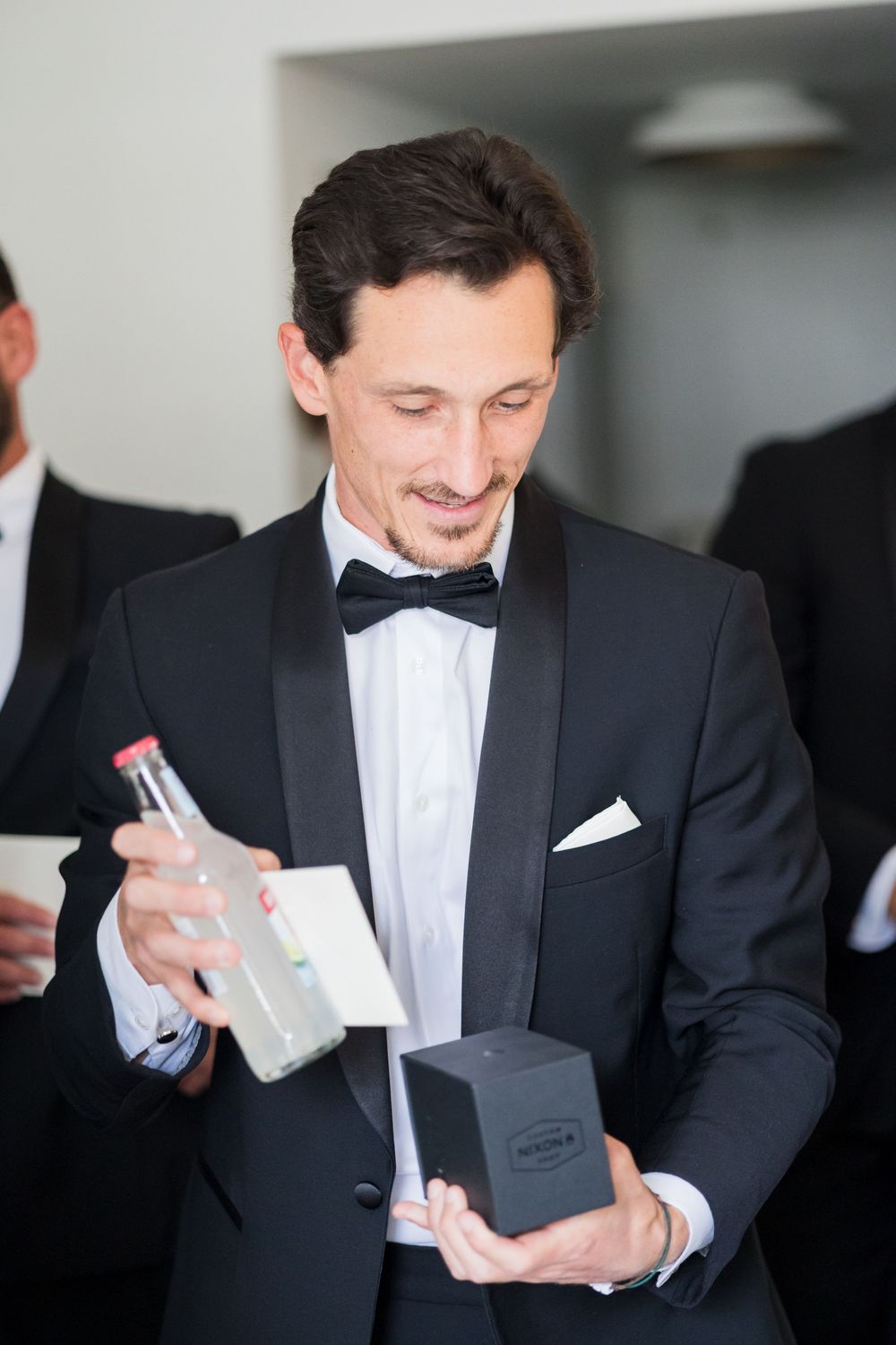 Elegant man in black tuxedo with bow tie holding a bottle and gift box at formal event.