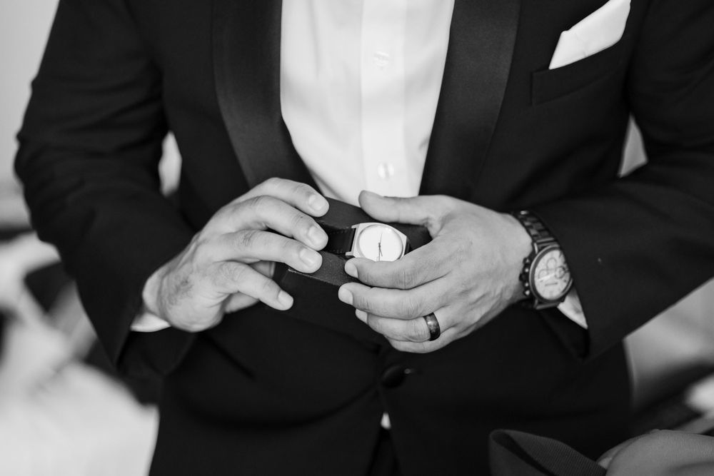 Groom in black tuxedo holds a watch while wearing a wedding band and wristwatch in black and white photo.