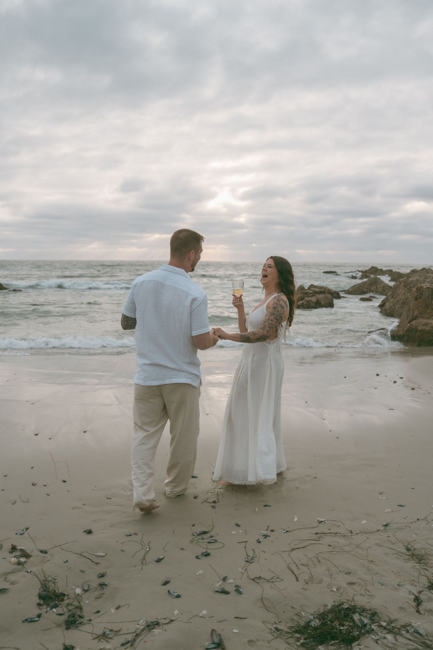 Couple in white attire holding champagne on a sandy beach at sunset with rocky shoreline and cloudy sky.