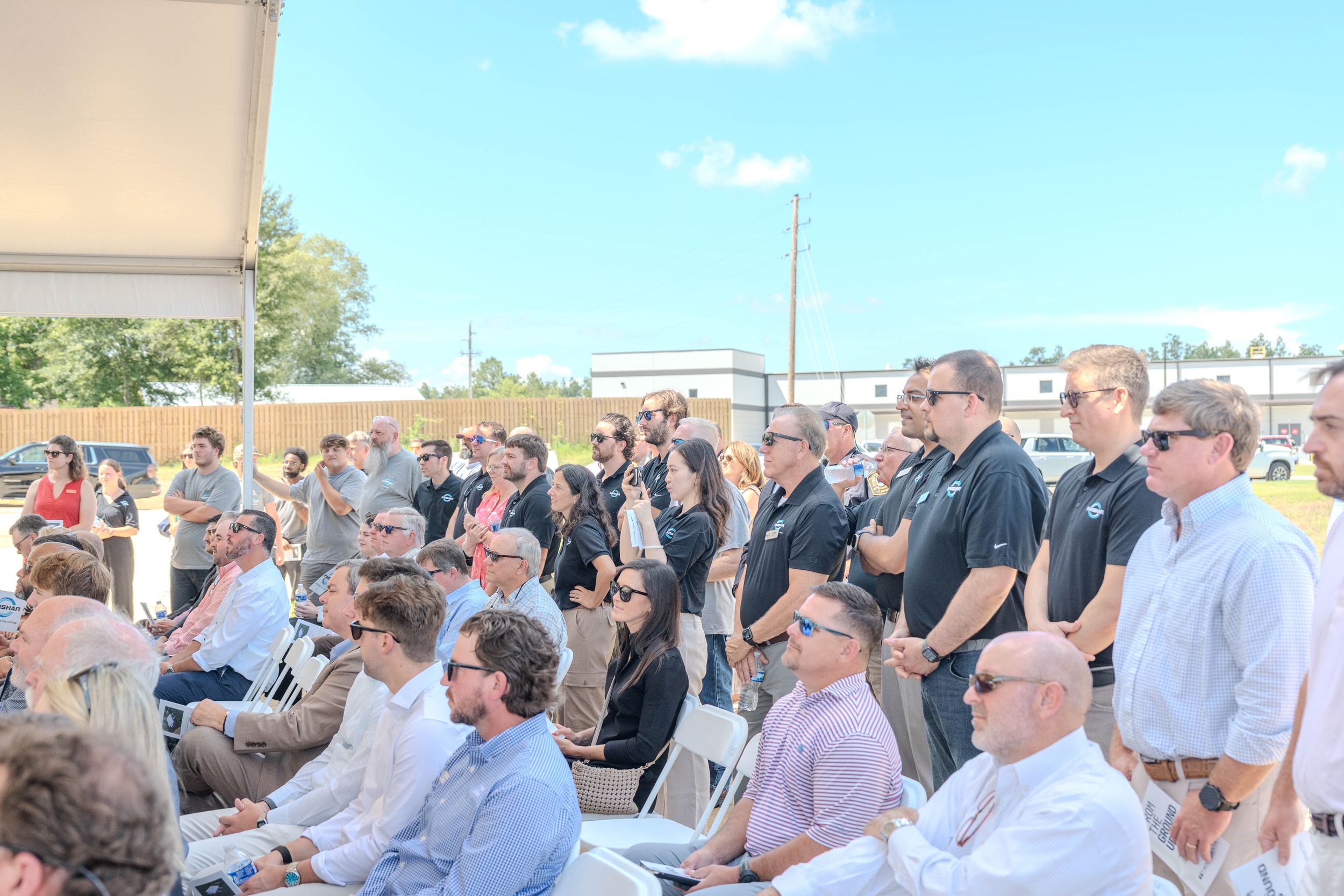Large group of professionals attending an outdoor corporate event or groundbreaking ceremony on a sunny day.