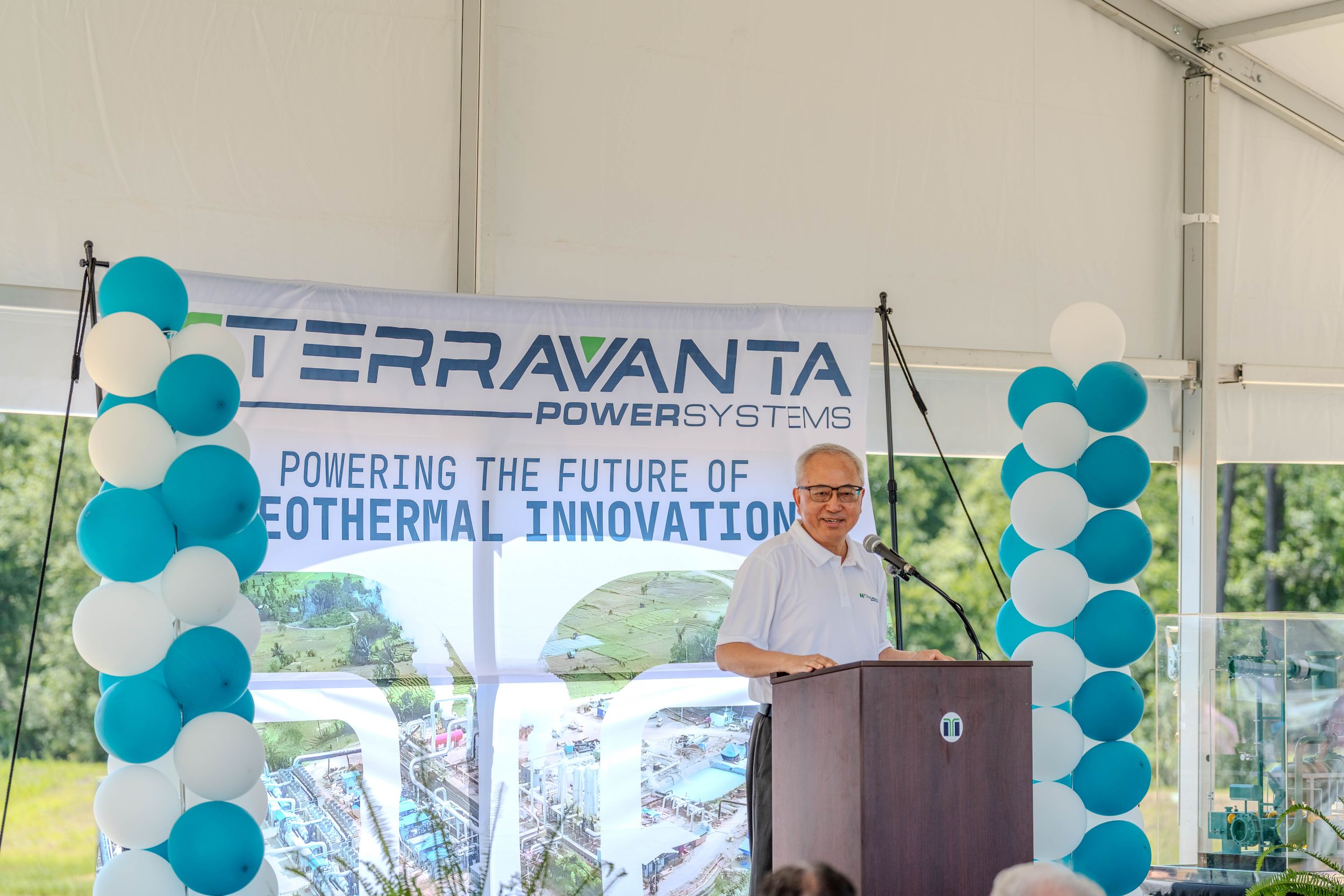 Speaker at Terravanta Power Systems podium promoting geothermal innovation at an outdoor event with teal balloon decorations.