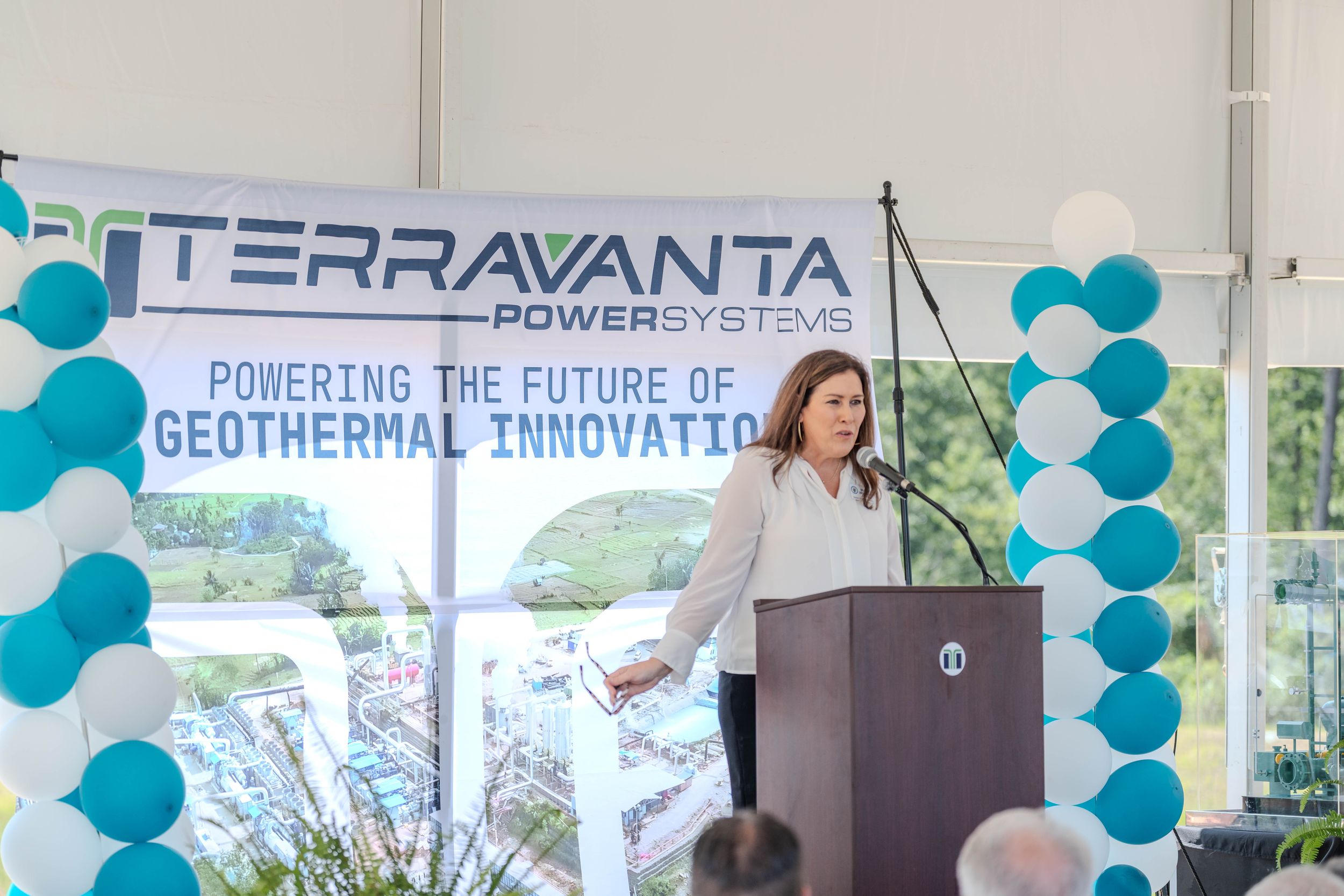 Woman speaking at Terravanta Power Systems geothermal innovation event podium with teal balloon decorations.