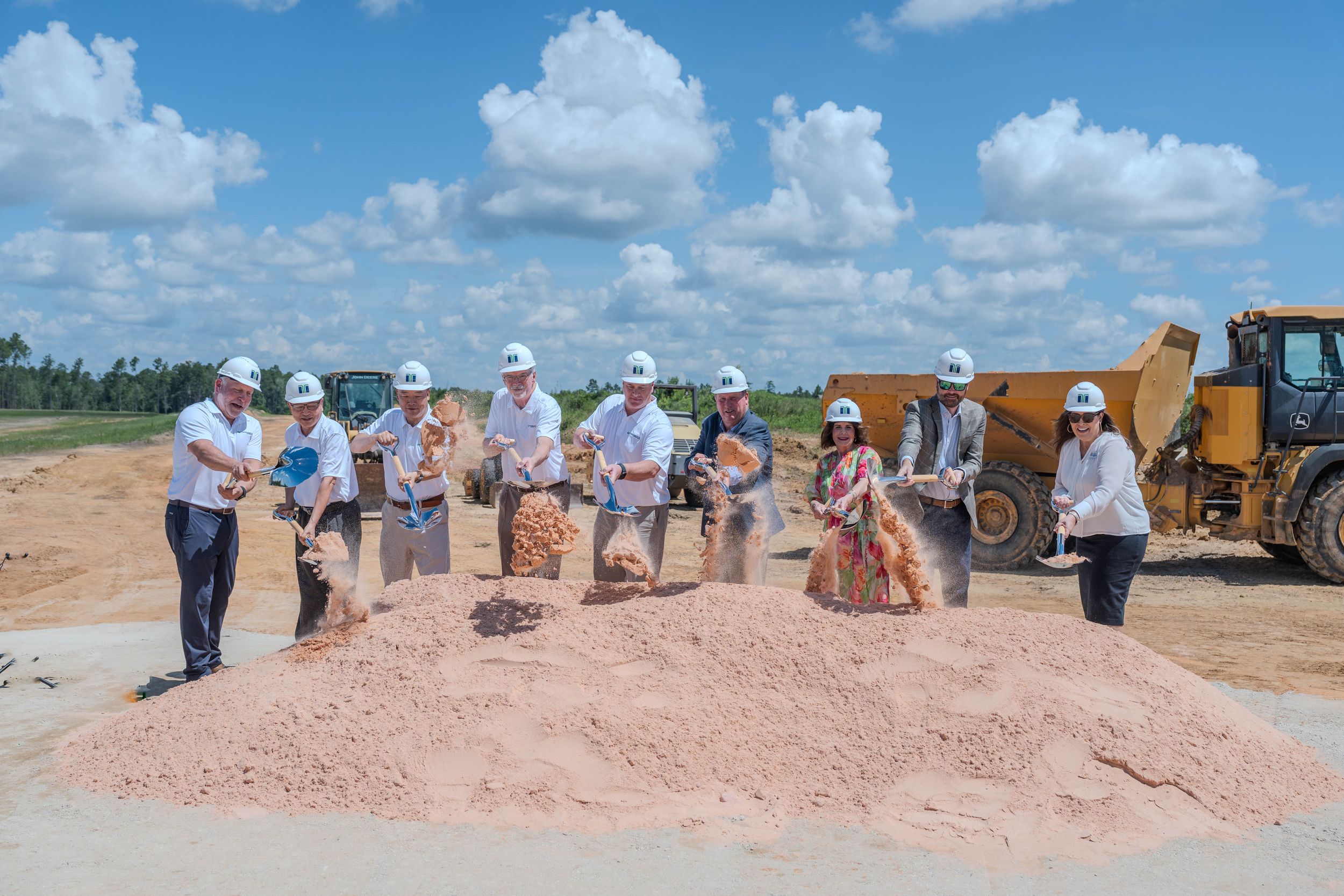 Group of executives in white hard hats ceremonially breaking ground at a construction site with shovels and a mound of dirt.
