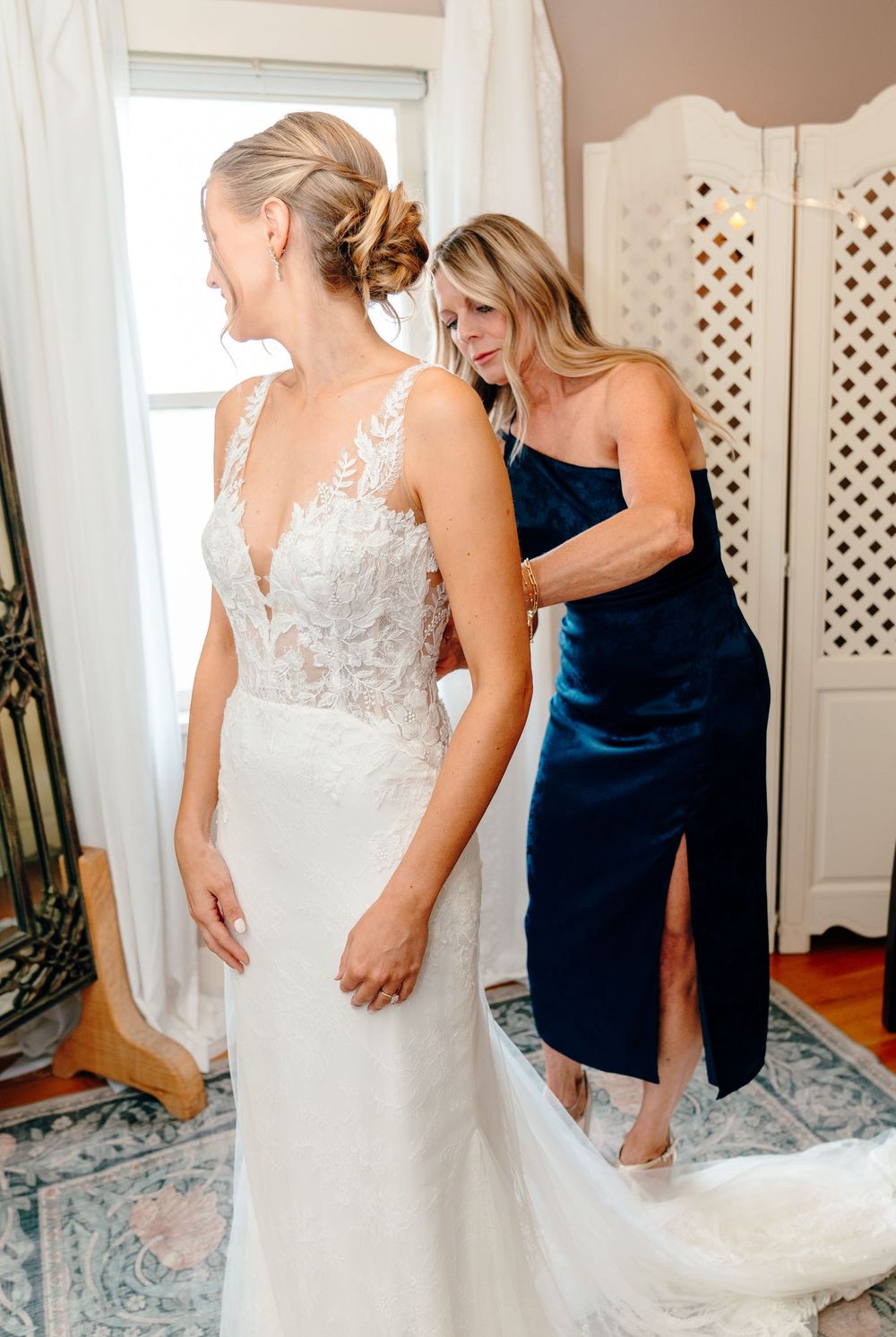 A bride in a lace wedding gown being helped by a woman in a navy dress in a bright, elegant room.