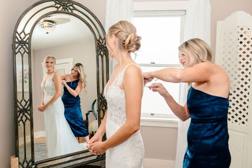 Bride in white lace gown getting dressed with bridesmaid in navy blue dress helping, reflected in ornate mirror.