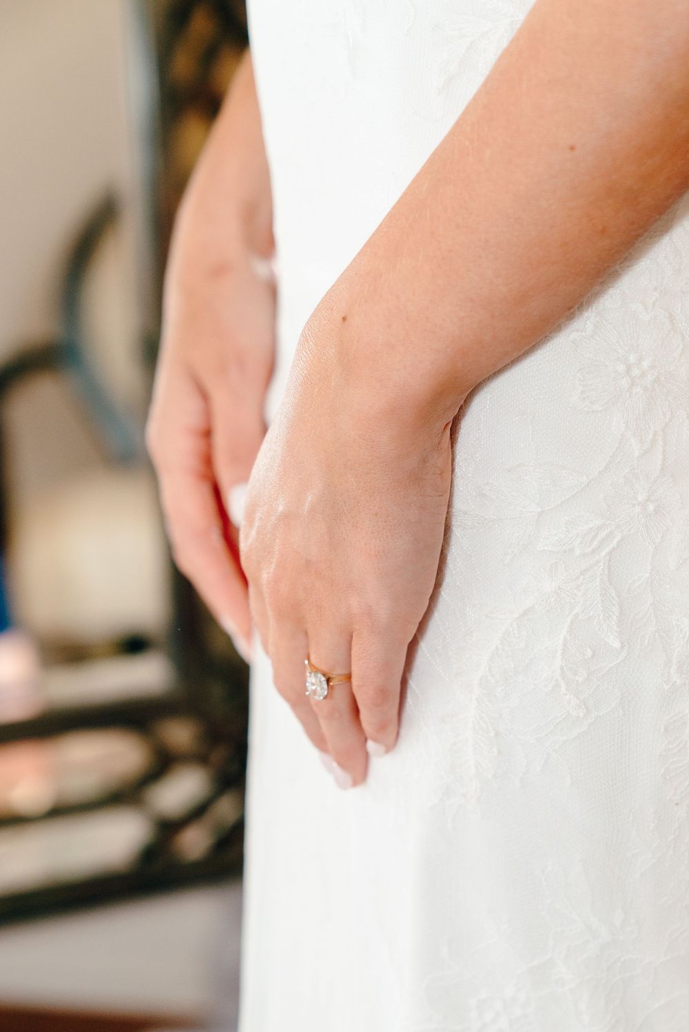 Close-up of a bride's hand wearing a gold solitaire diamond engagement ring against her white lace wedding dress.