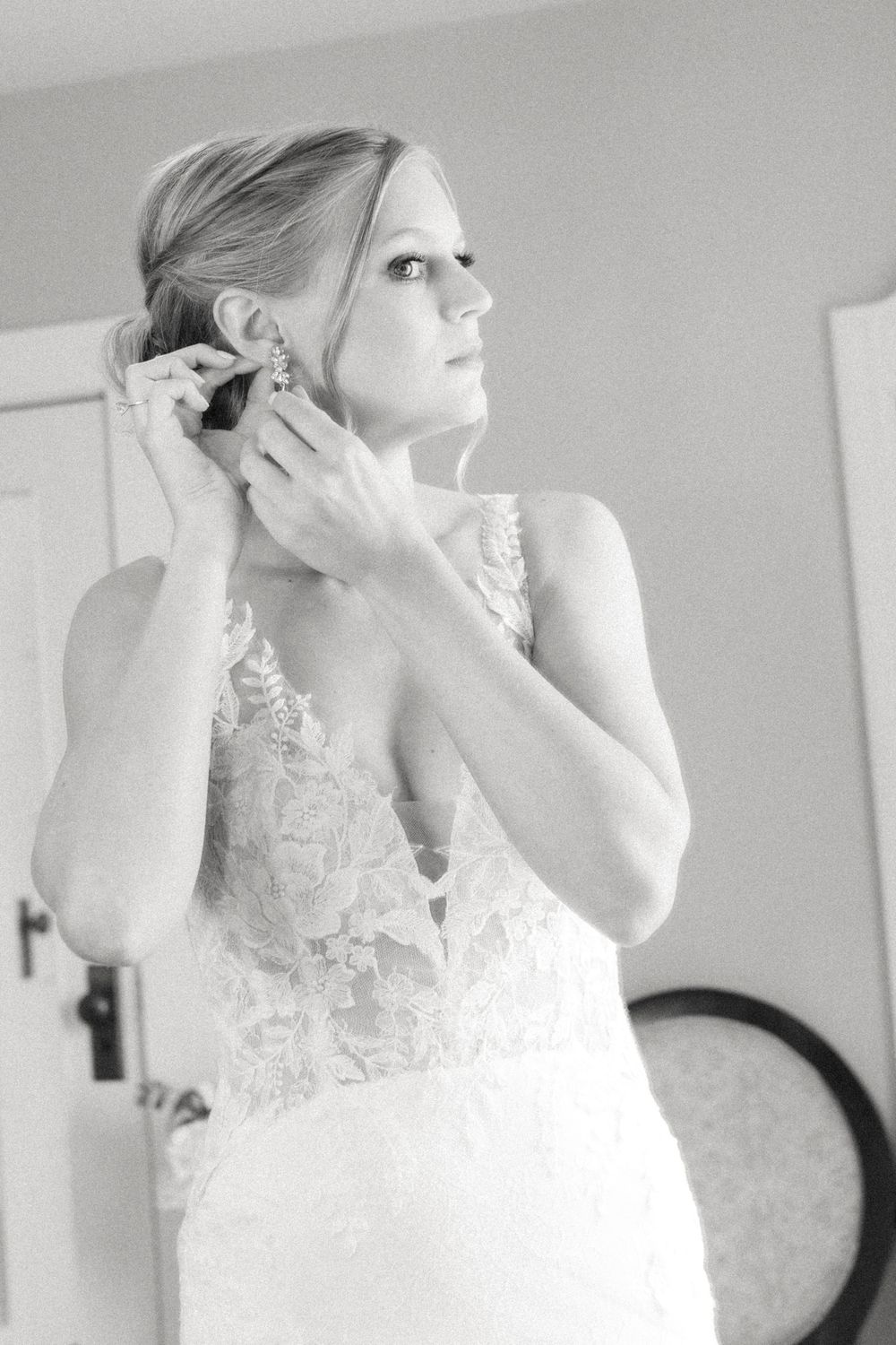 Bride in lace wedding gown putting on earrings in black and white photo.