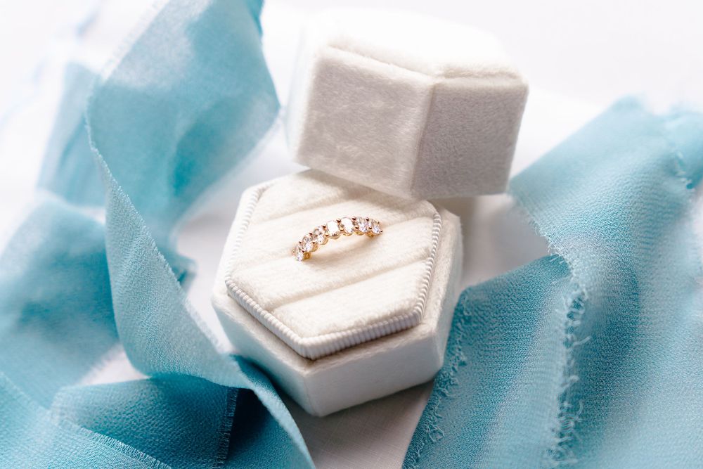 Gold diamond ring displayed in a white velvet hexagonal ring box with soft blue silk ribbon backdrop.
