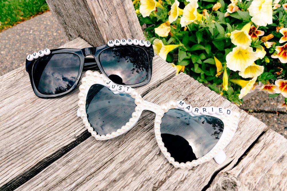 Two decorative sunglasses with pearl trim on a wooden surface, one black and one white heart-shaped reading Just Married.