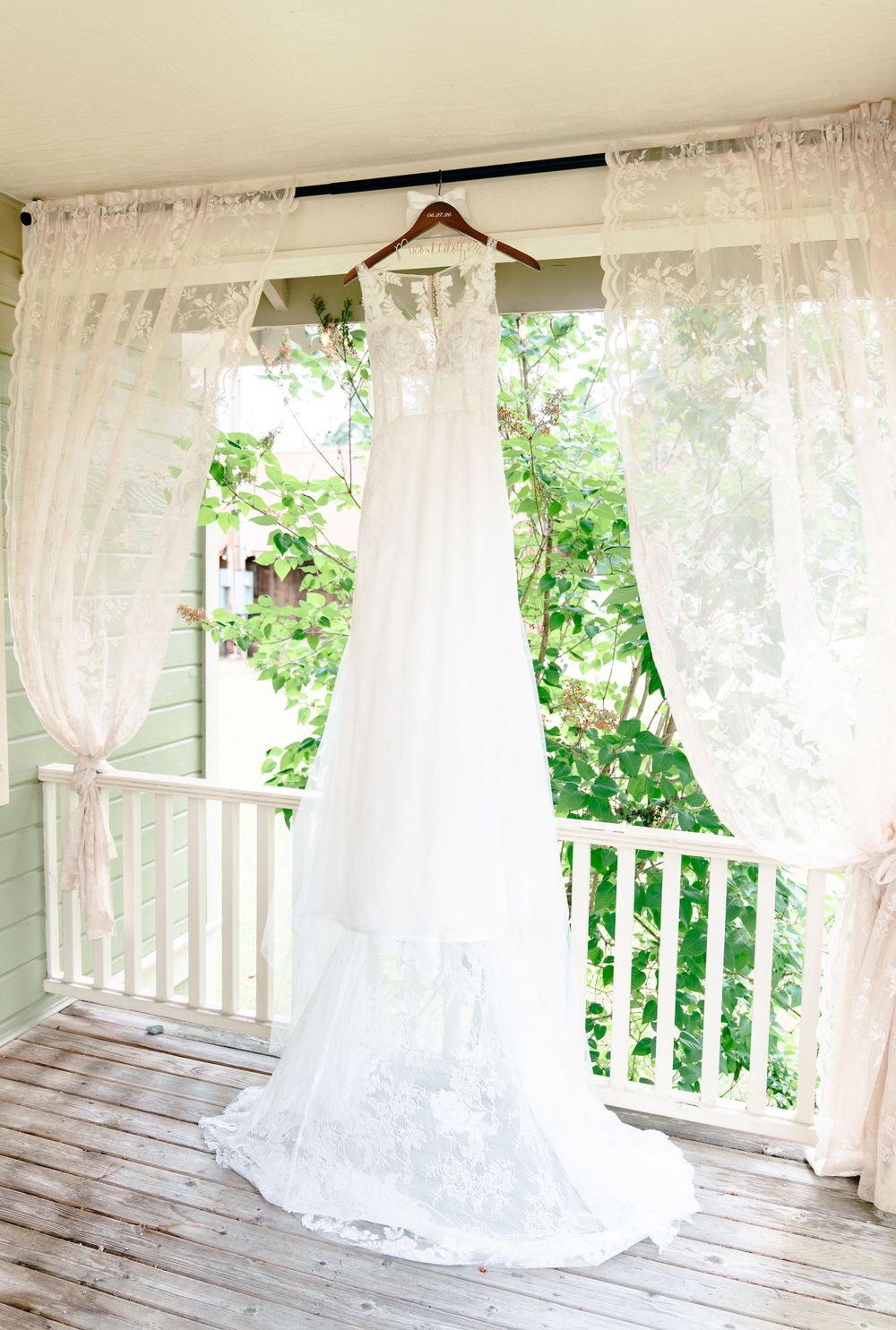 White lace wedding dress hanging on a wooden hanger between sheer curtains on a charming outdoor porch.