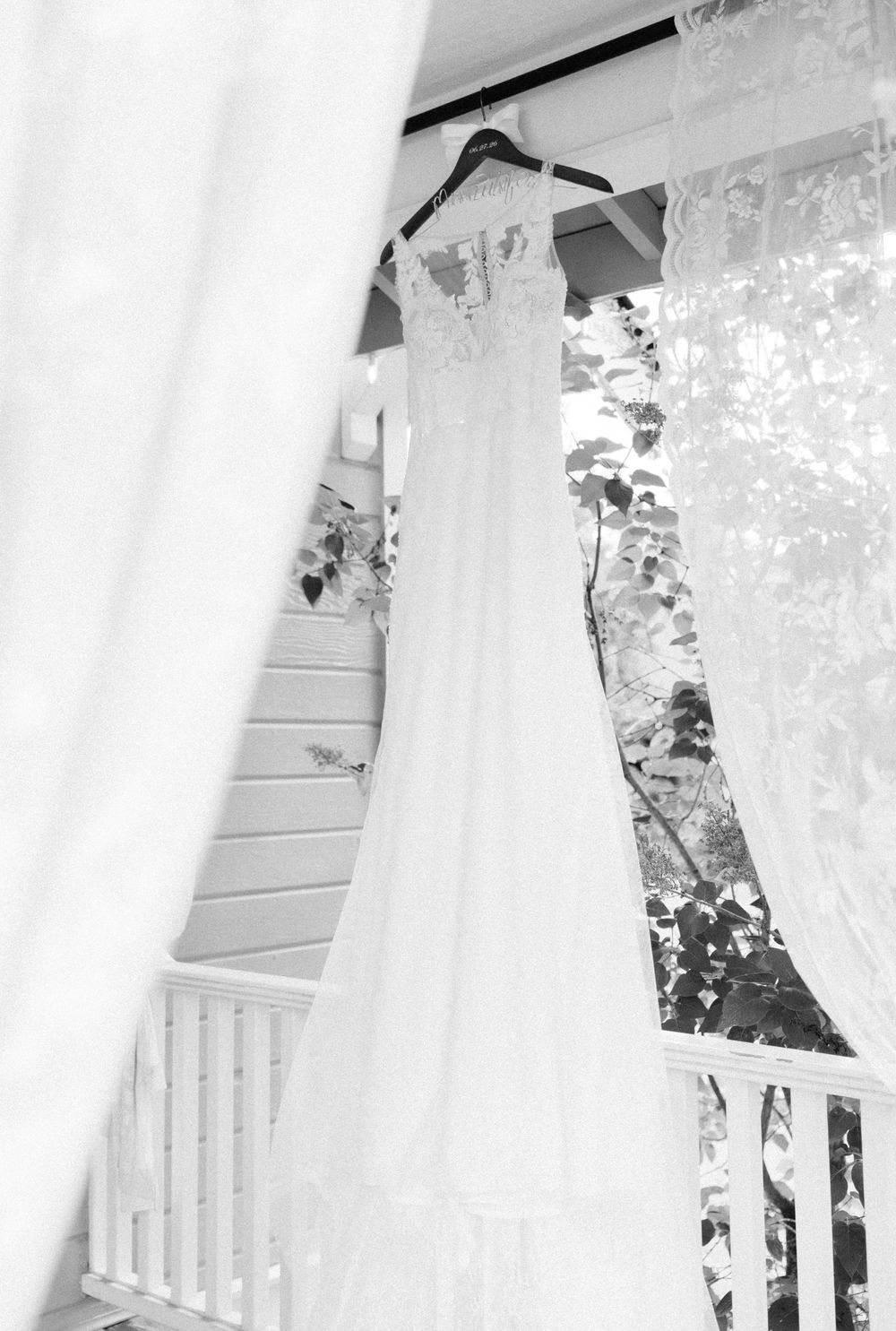 Black and white photo of an elegant lace wedding dress hanging on a porch hanger with sheer curtains and greenery.