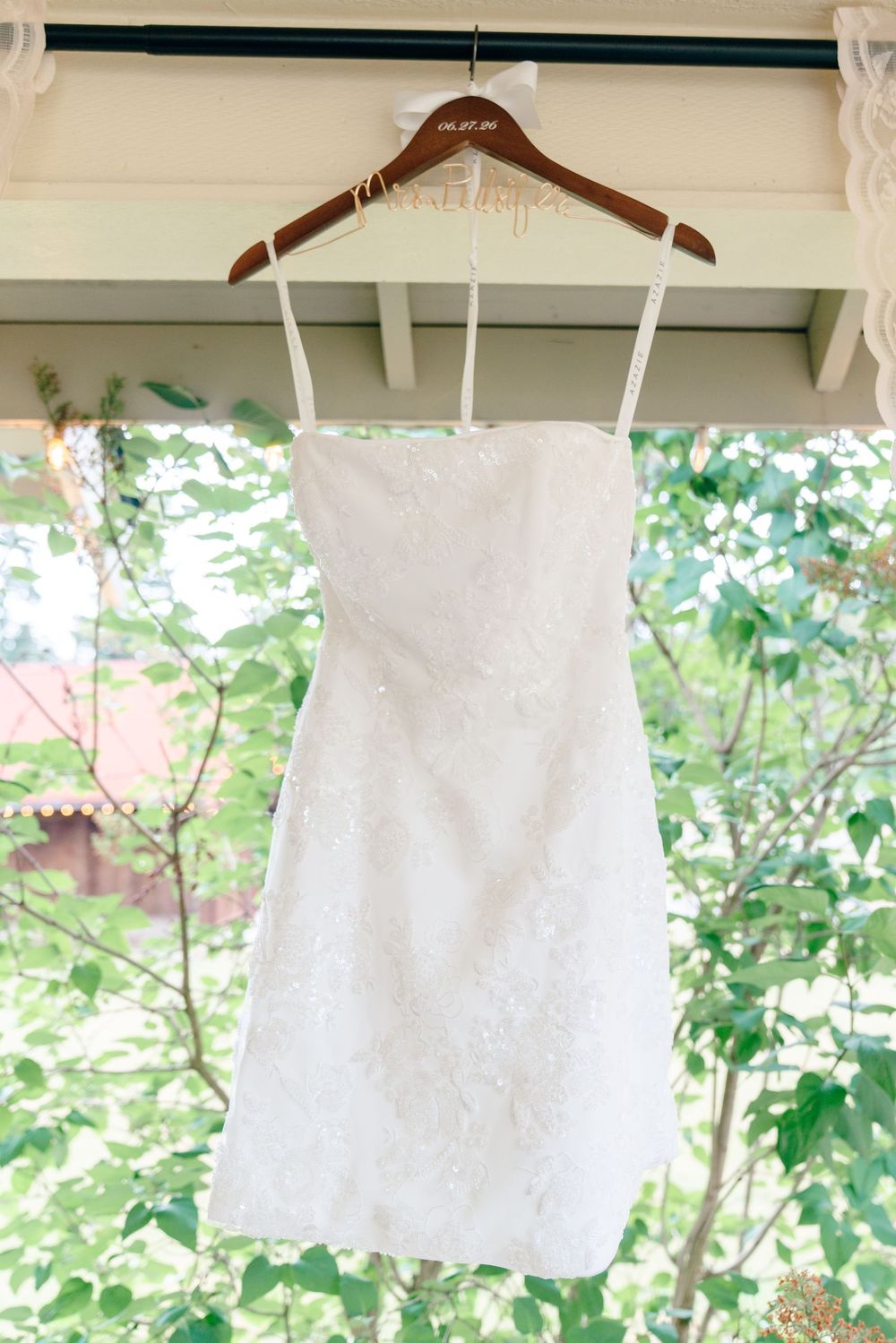 White lace mini wedding dress on wooden hanger displayed outdoors with green foliage background.