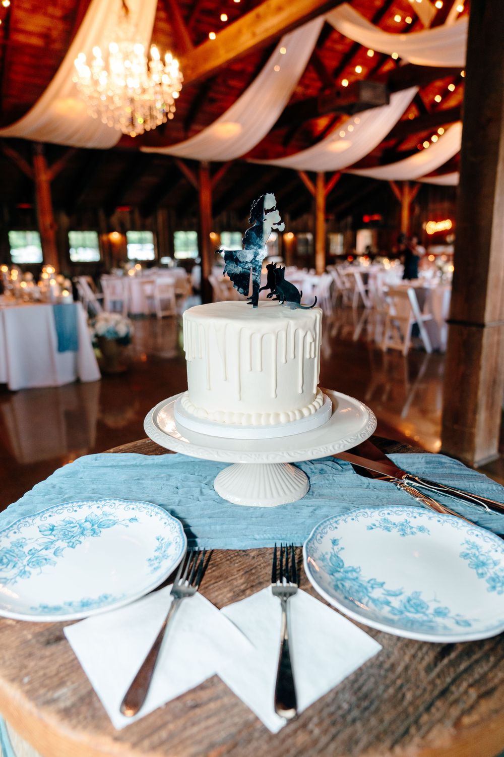 White single-tier wedding cake with figurine topper on blue runner in rustic barn reception venue with chandeliers.