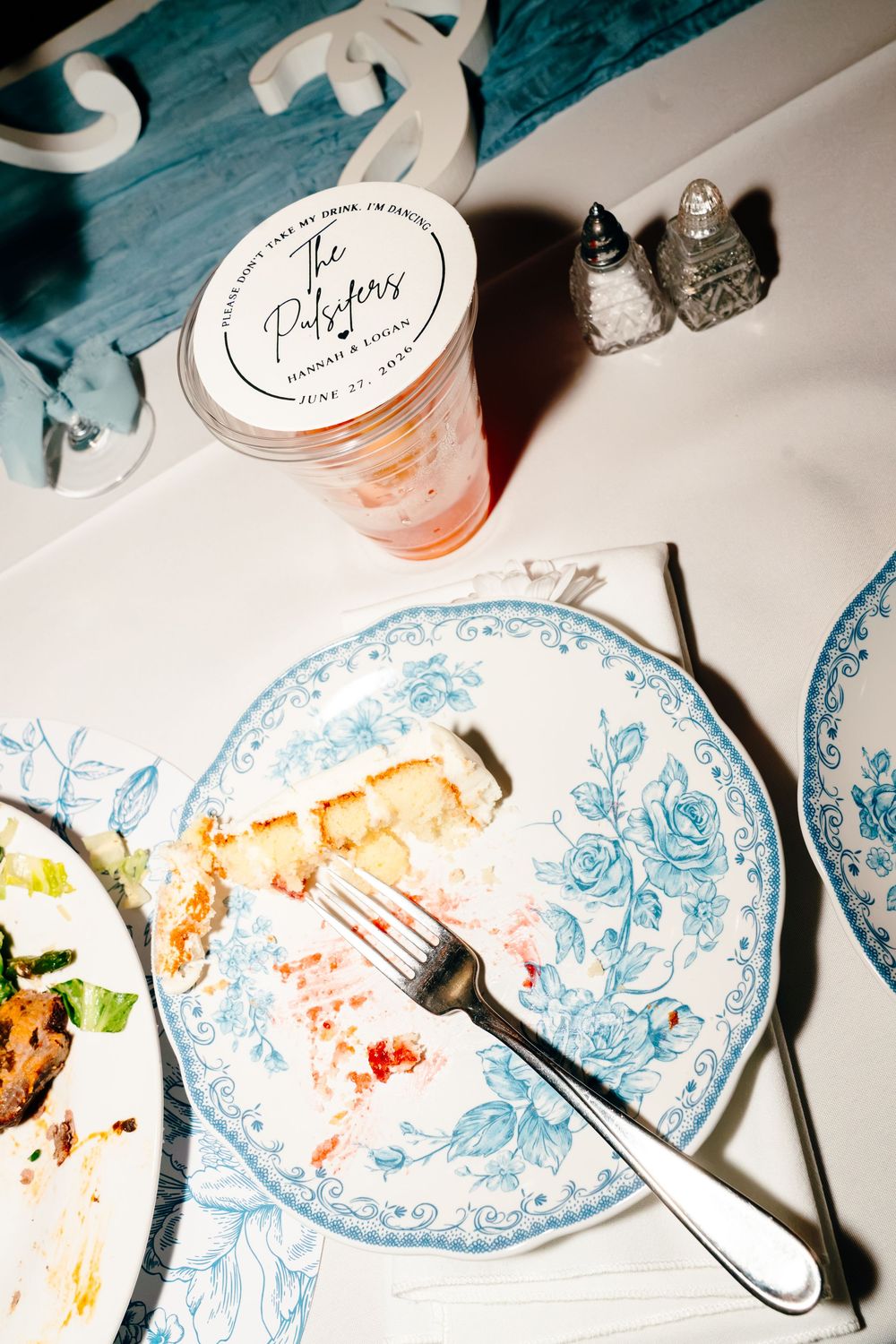 Blue floral plate with wedding cake slice and fork beside personalized couples cup at reception table.