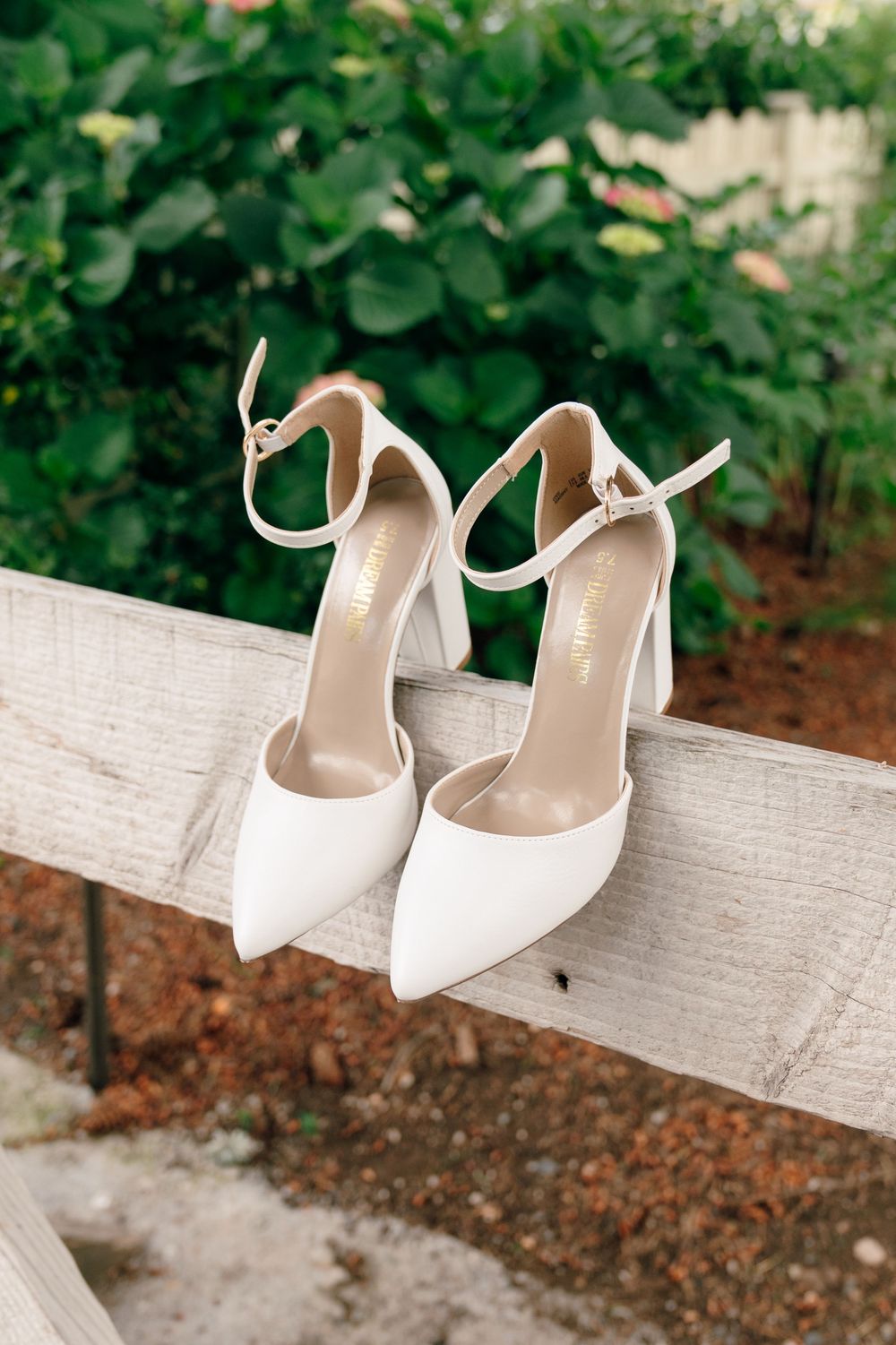 Elegant white pointed-toe heels with ankle straps displayed on a wooden fence with lush green foliage backdrop.