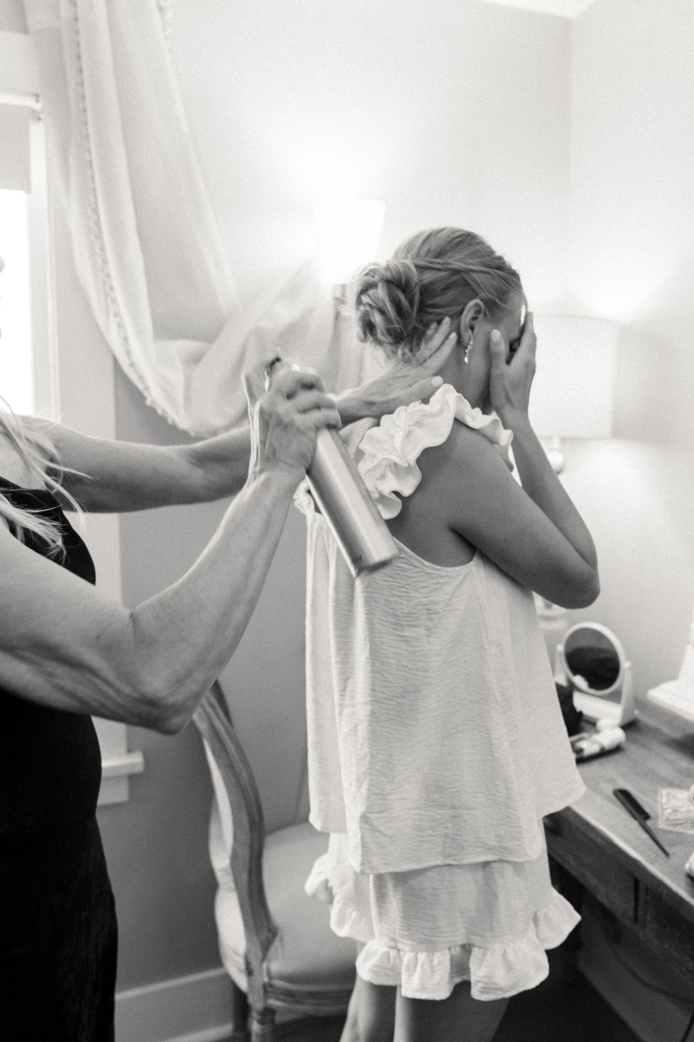 Black and white photo of a bride getting hair sprayed while covering her face during wedding preparations.
