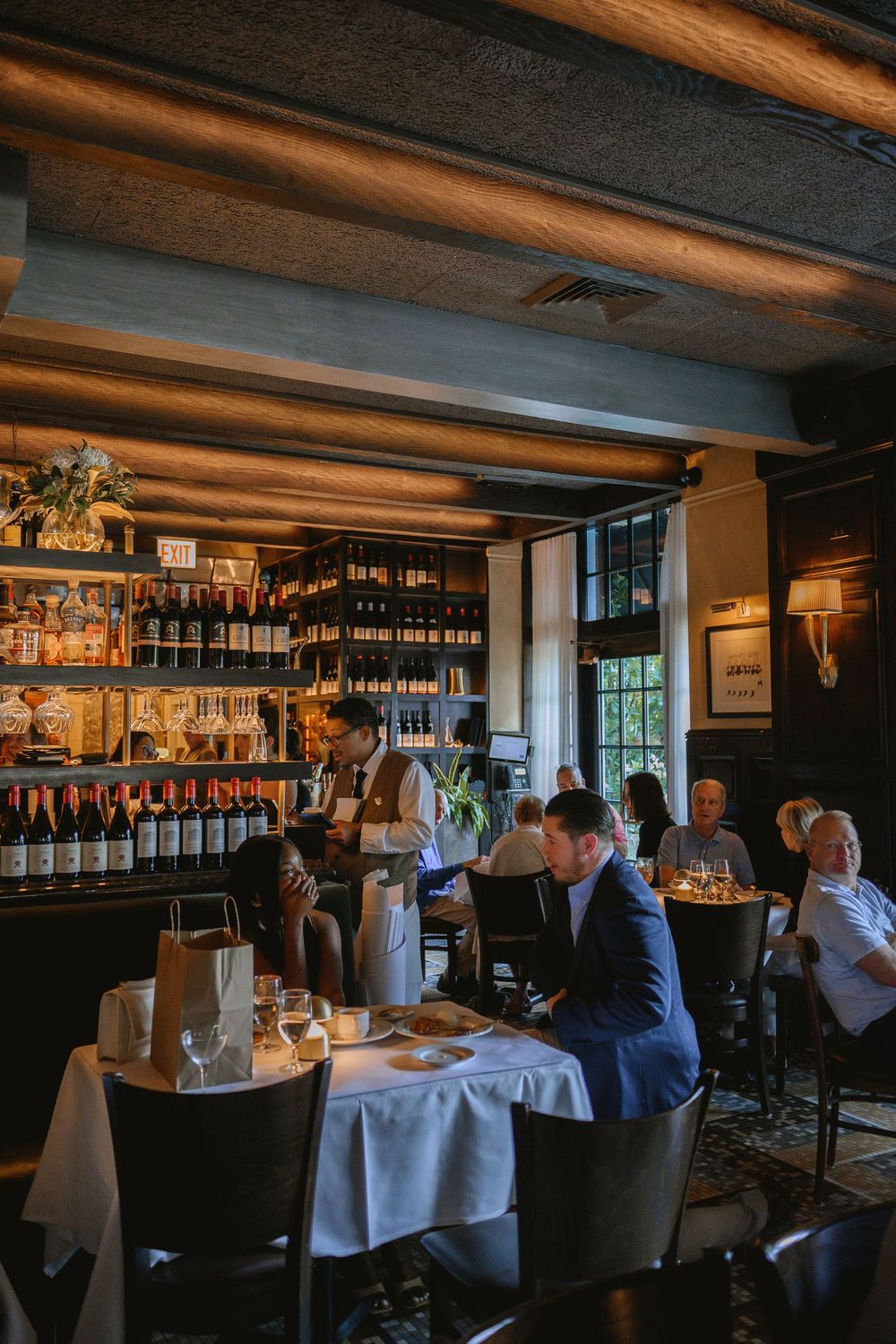 Dimly lit upscale restaurant interior with wine shelves, white tablecloths, and elegantly dressed diners enjoying an evening meal.