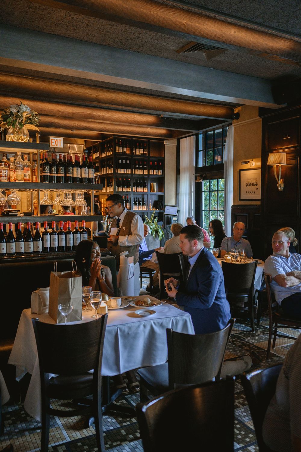 Elegant restaurant interior with wine bar, white tablecloths, and diners enjoying meals in warm ambient lighting.