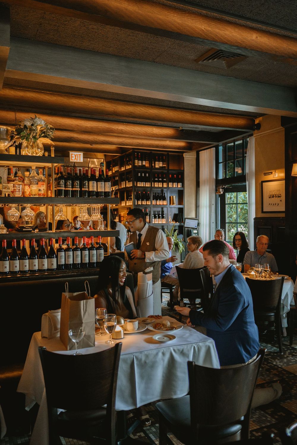 Elegant restaurant interior with wine shelves, white tablecloths, and diners enjoying meals in warm ambient lighting.