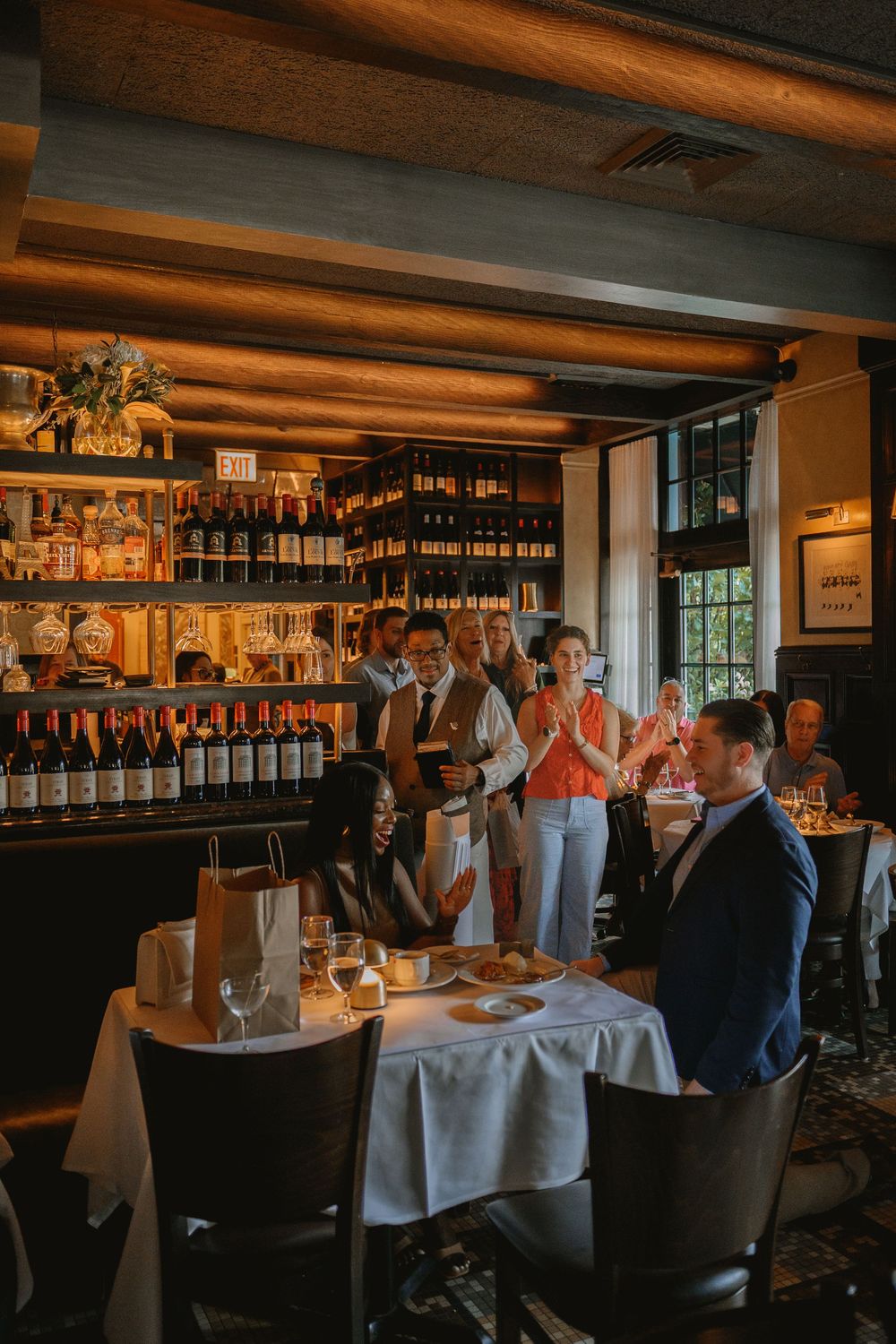 Elegant restaurant dining scene with guests at candlelit table, wine wall backdrop, and staff serving in warm ambient lighting.