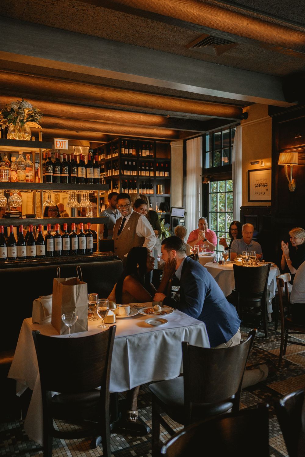 Elegant restaurant interior with wine bar, diners at white tablecloth tables, and warm ambient lighting.