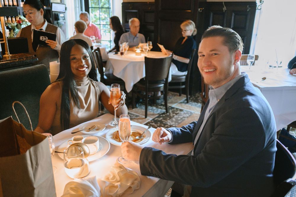 Couple smiling at dinner table in elegant restaurant, enjoying drinks and appetizers in a bright dining room.