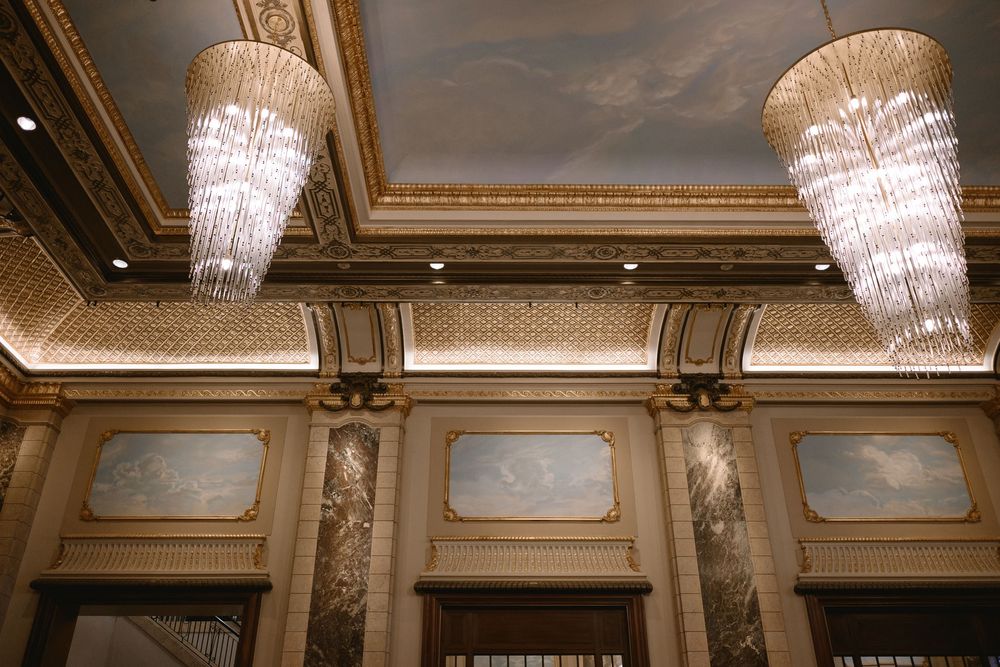 Elegant ballroom ceiling with ornate gold molding, two crystal chandeliers, and classical mural wall panels.