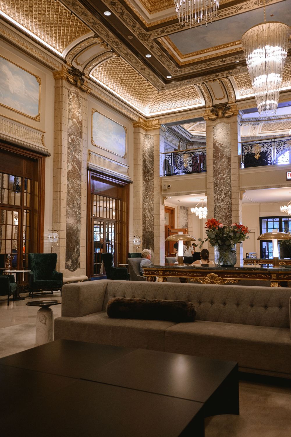 Grand hotel lobby featuring marble columns, ornate gold ceiling, crystal chandeliers, and plush gray sofas in a luxurious setting.