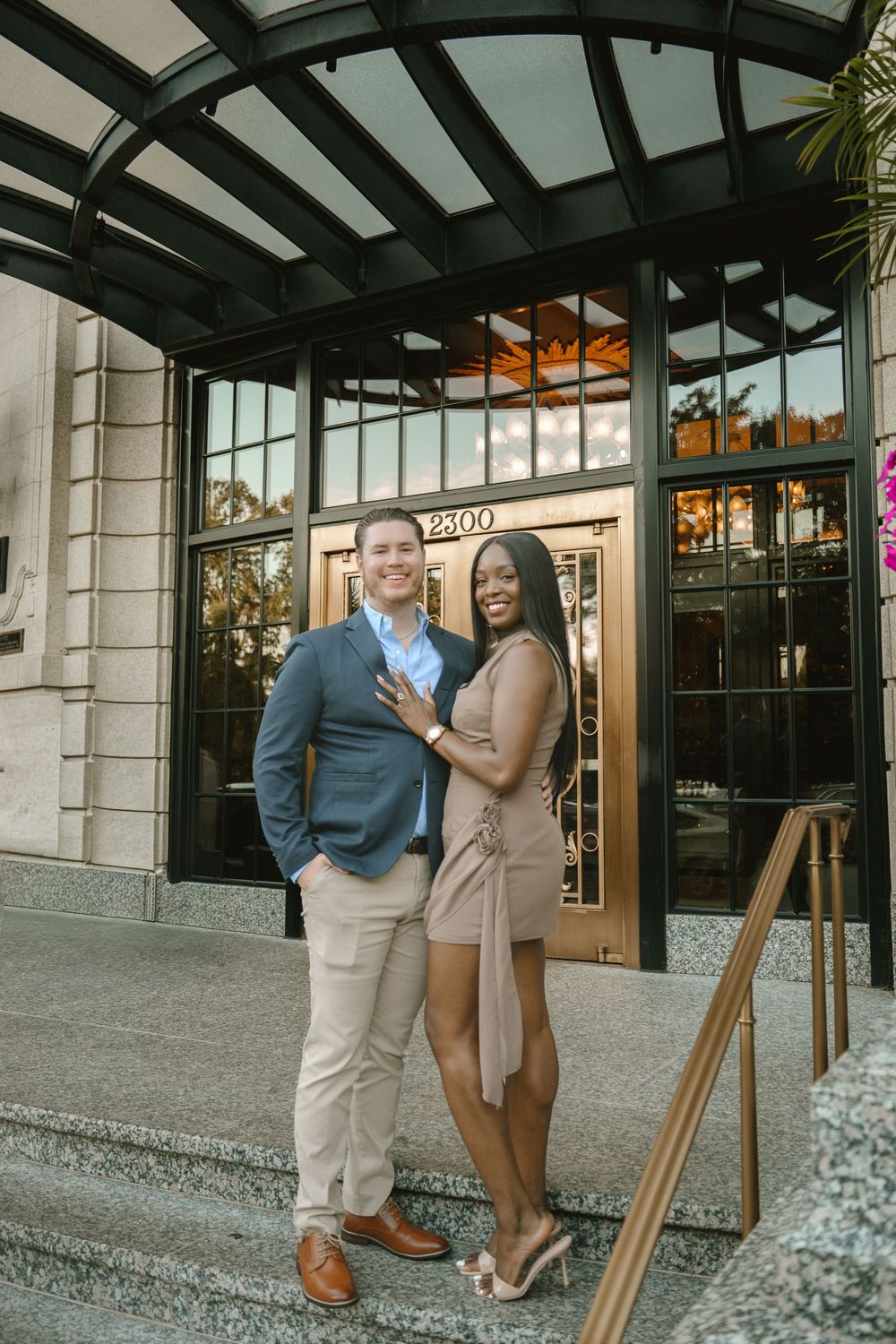 Smiling interracial couple posing together outside an elegant building entrance with address number 2300 and glass doors.