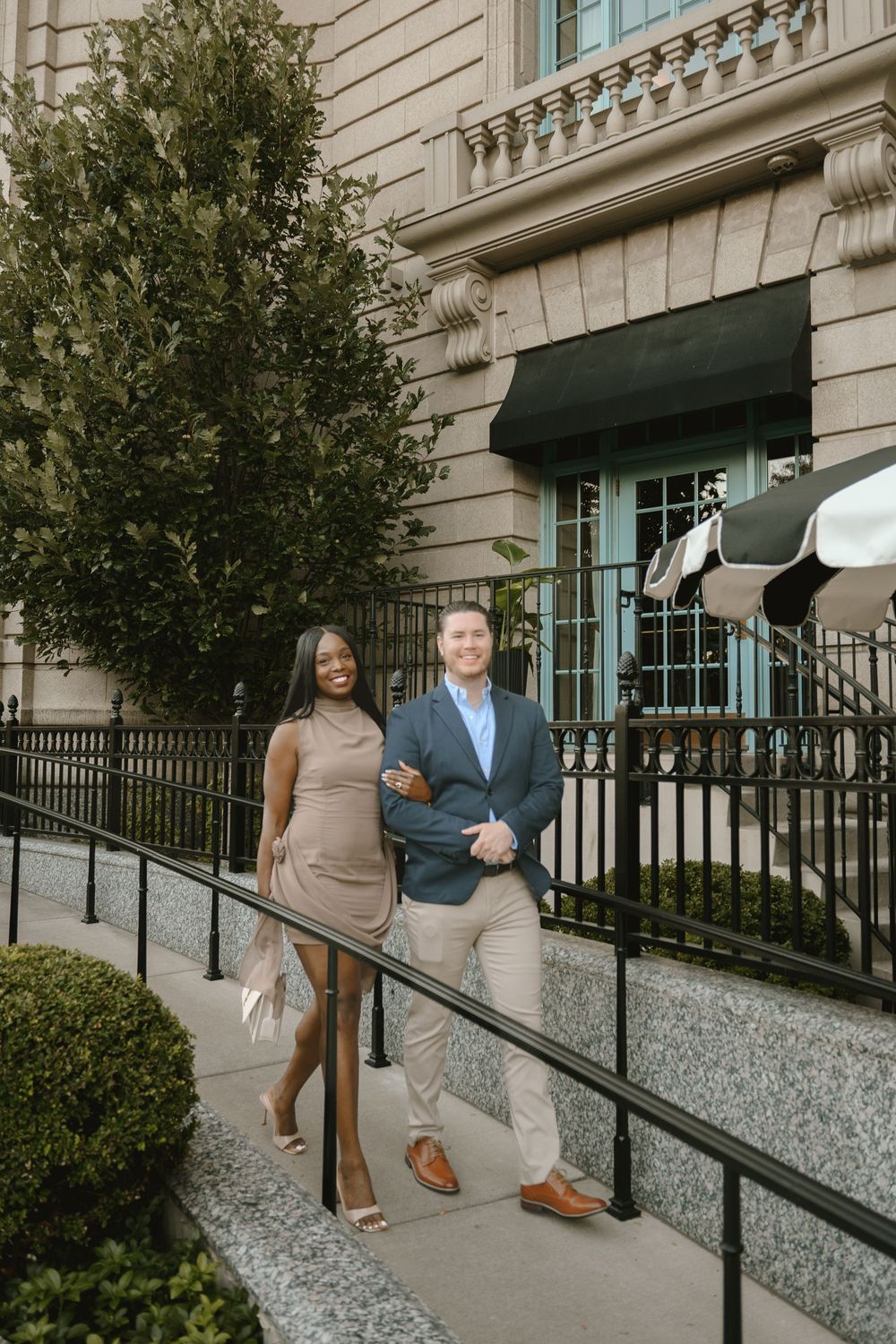 A smiling couple poses on steps outside an elegant building, woman in tan dress, man in blue blazer and khakis.