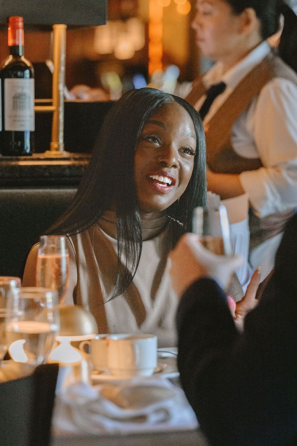 A smiling Black woman enjoys a fine dining experience at a restaurant table with glasses and candles.