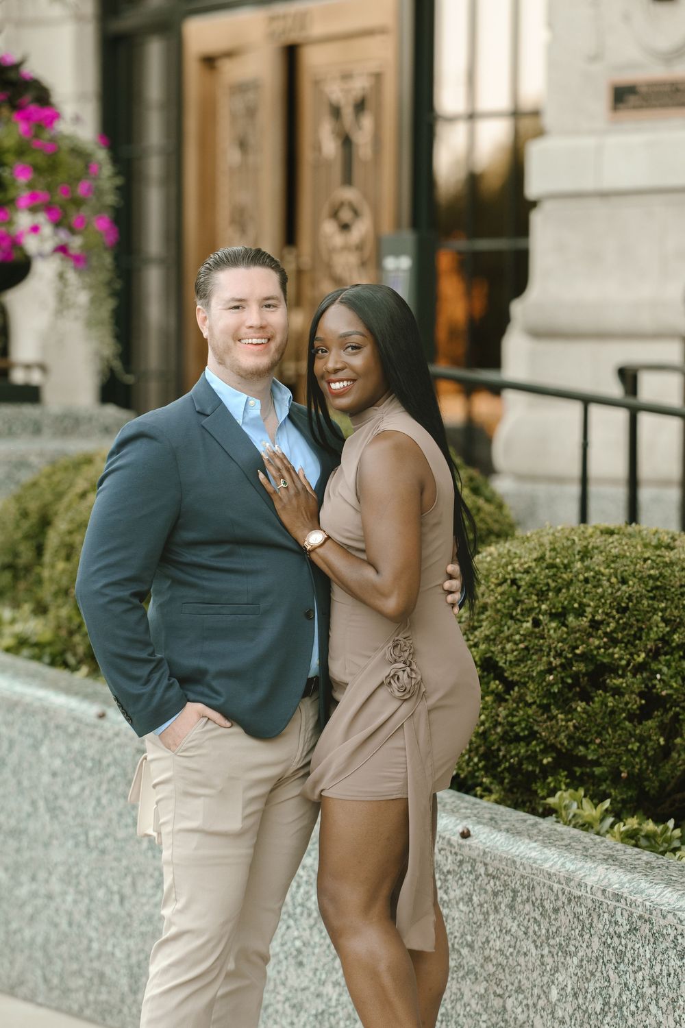 A smiling interracial couple poses elegantly outside a building, woman showing engagement ring, wearing smart casual attire.