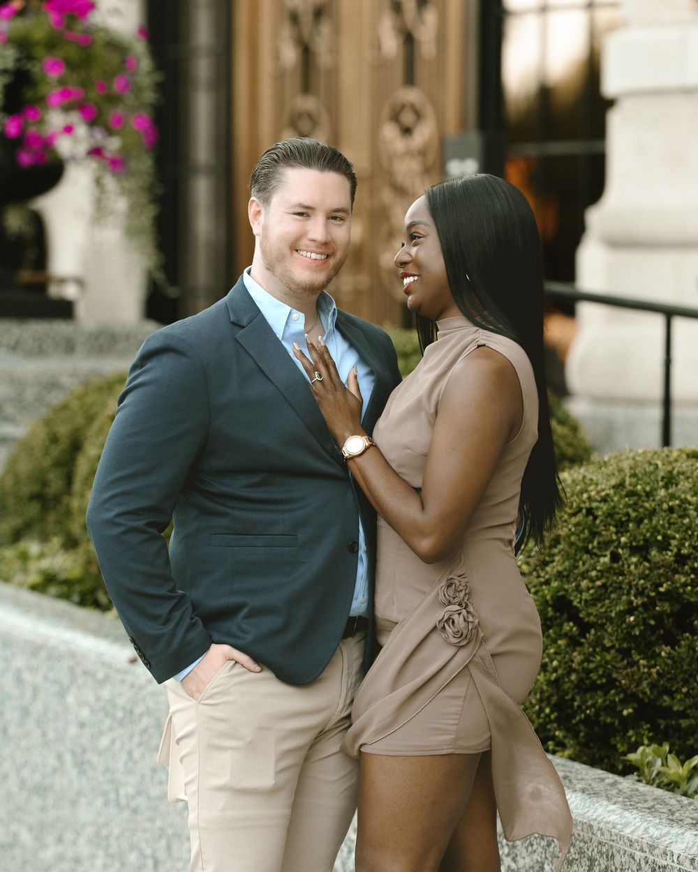 A smiling couple poses outdoors, man in navy blazer and woman in tan dress, sharing a joyful moment near elegant architecture.