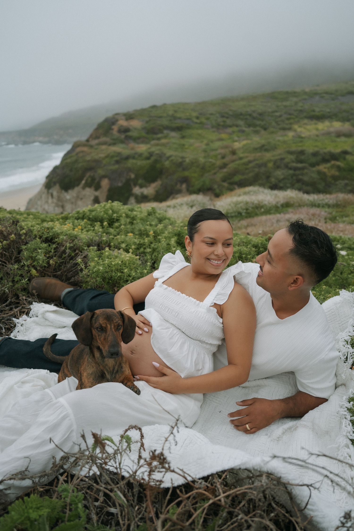 Couple relaxing on white blanket outdoors with dachshund dog, coastal cliffs and ocean in misty background.