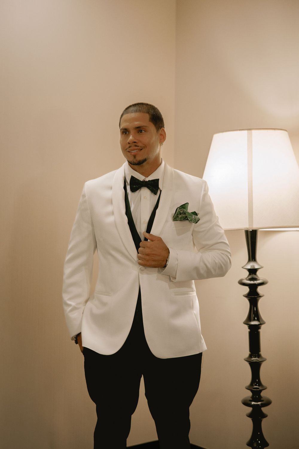 Groom in white tuxedo jacket with black bow tie and green pocket square posing elegantly beside a floor lamp.