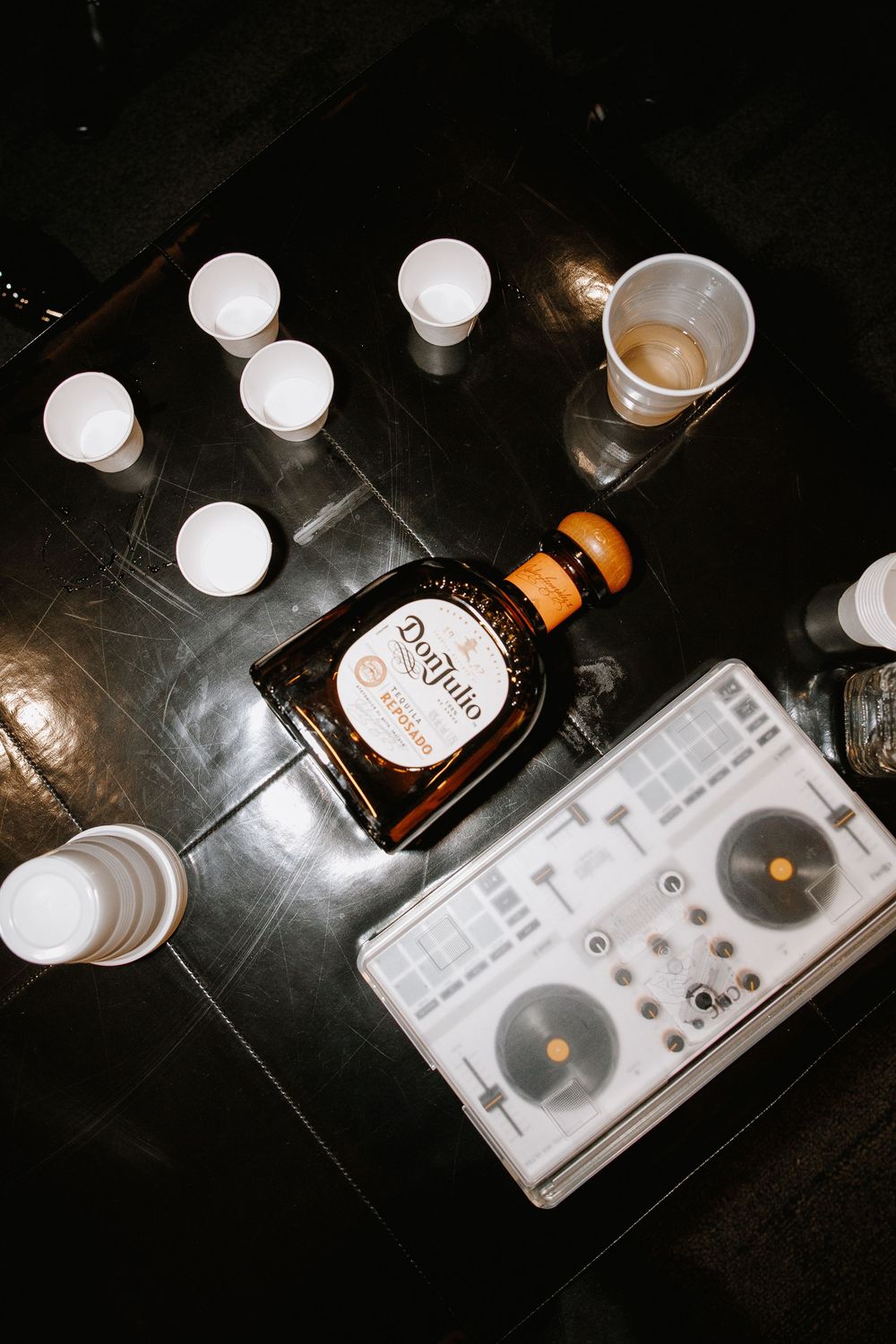 Don Julio tequila bottle beside a DJ controller, surrounded by white cups on a dark table.
