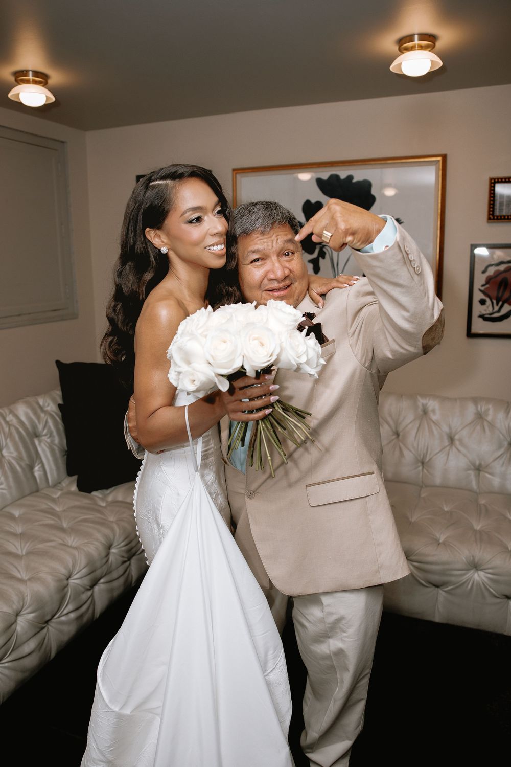Bride in white gown holds bouquet of white roses while posing with older man in beige suit at wedding.