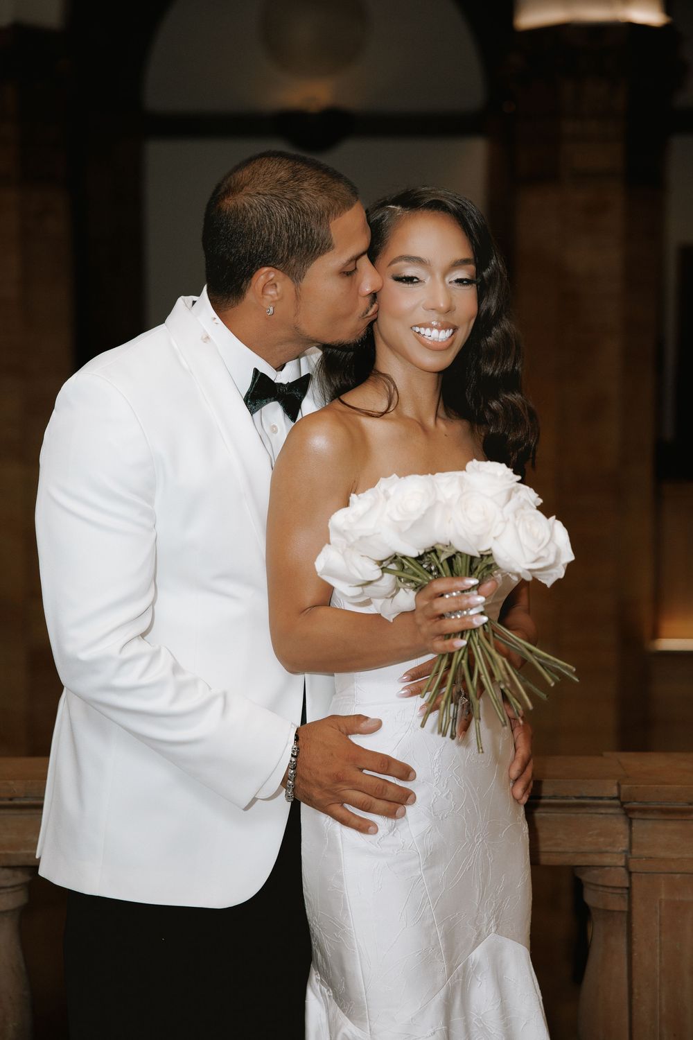 Bride and groom in white wedding attire share a romantic kiss, bride holding white rose bouquet.