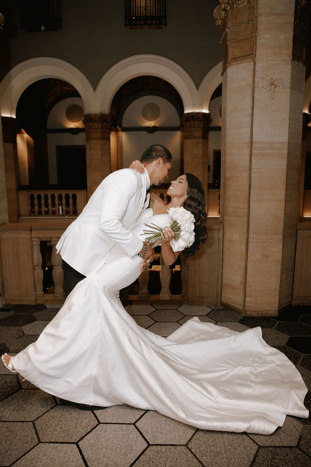 Bride and groom share a romantic dip kiss, she in a white mermaid gown, he in a white tuxedo, in an elegant venue.