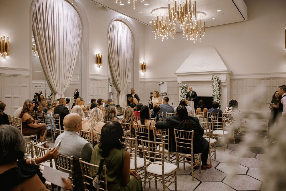 Elegant wedding ceremony in a grand ballroom with gold chairs, chandeliers, and draped white curtains.