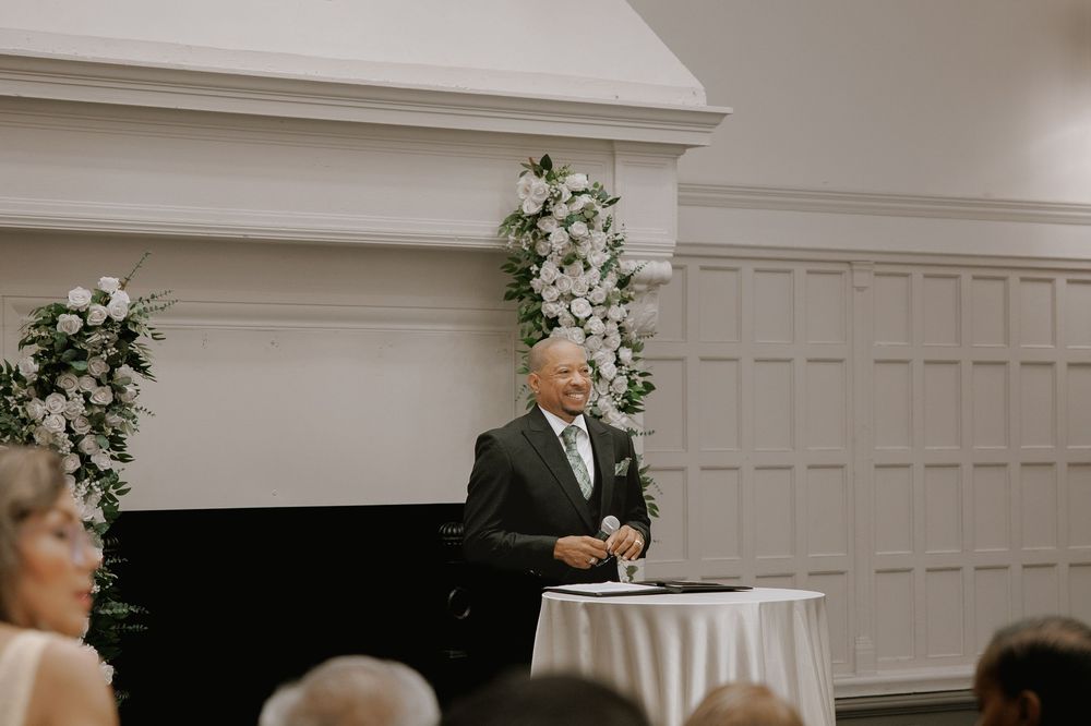 Officiant smiling at wedding ceremony podium with white floral arrangements flanking elegant fireplace backdrop.