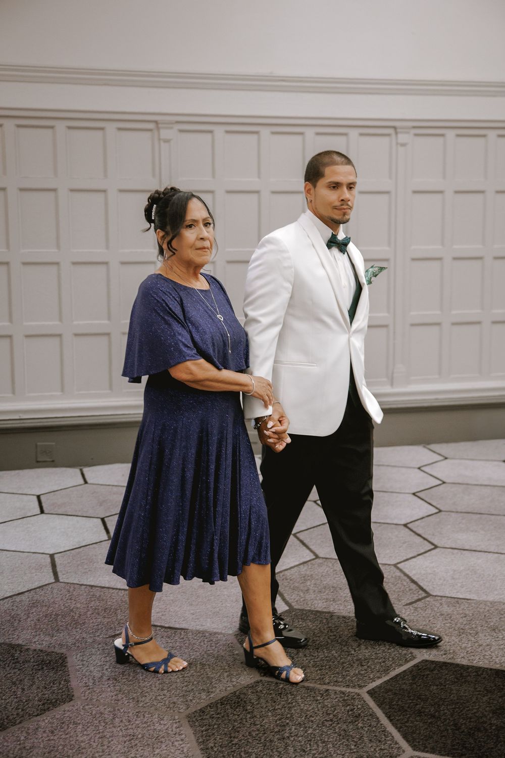 A young man in a white tuxedo jacket escorts an older woman in a navy blue dress at a formal wedding event.
