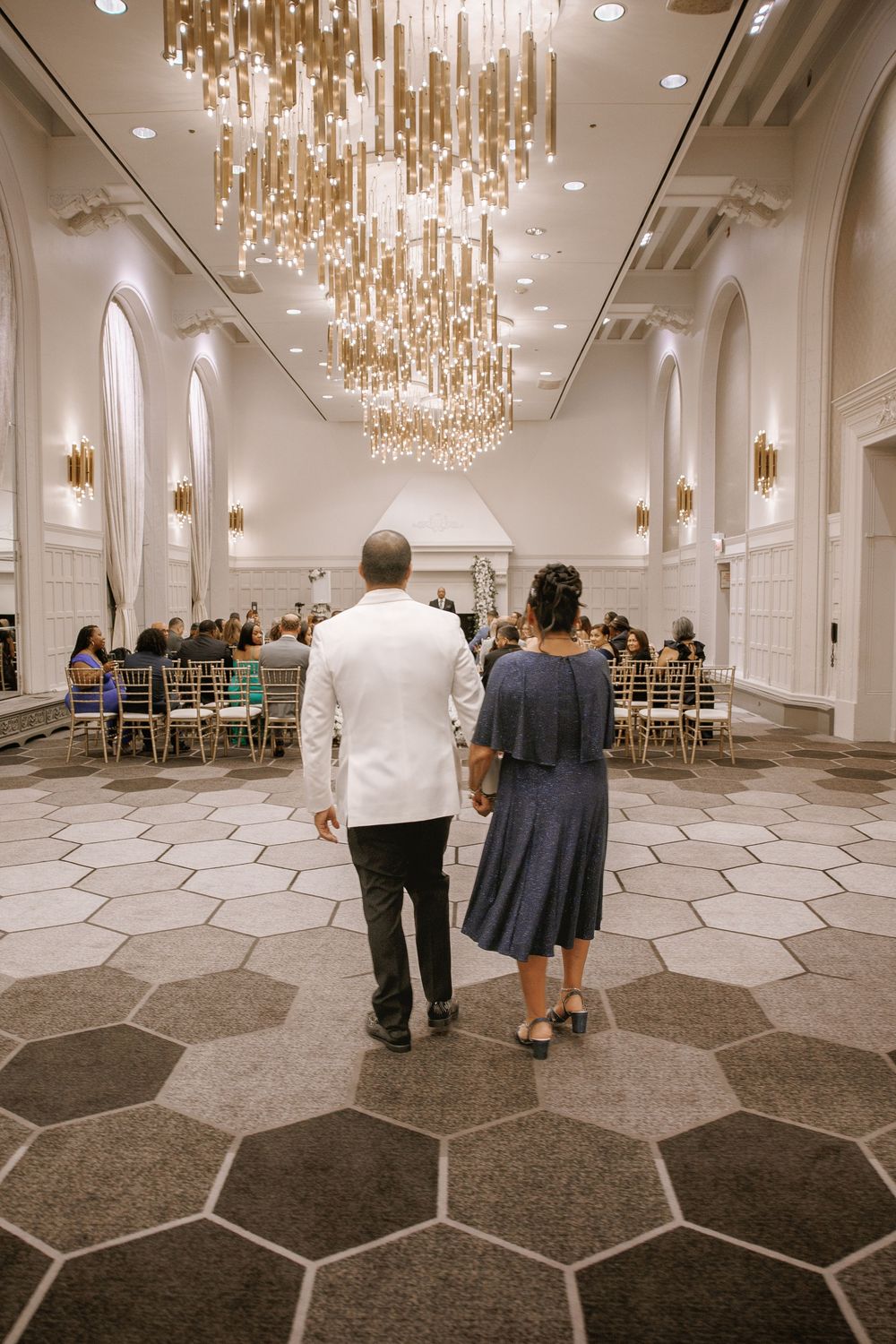 Couple walks hand-in-hand toward wedding ceremony in elegant ballroom with hexagonal floors and gold chandelier.