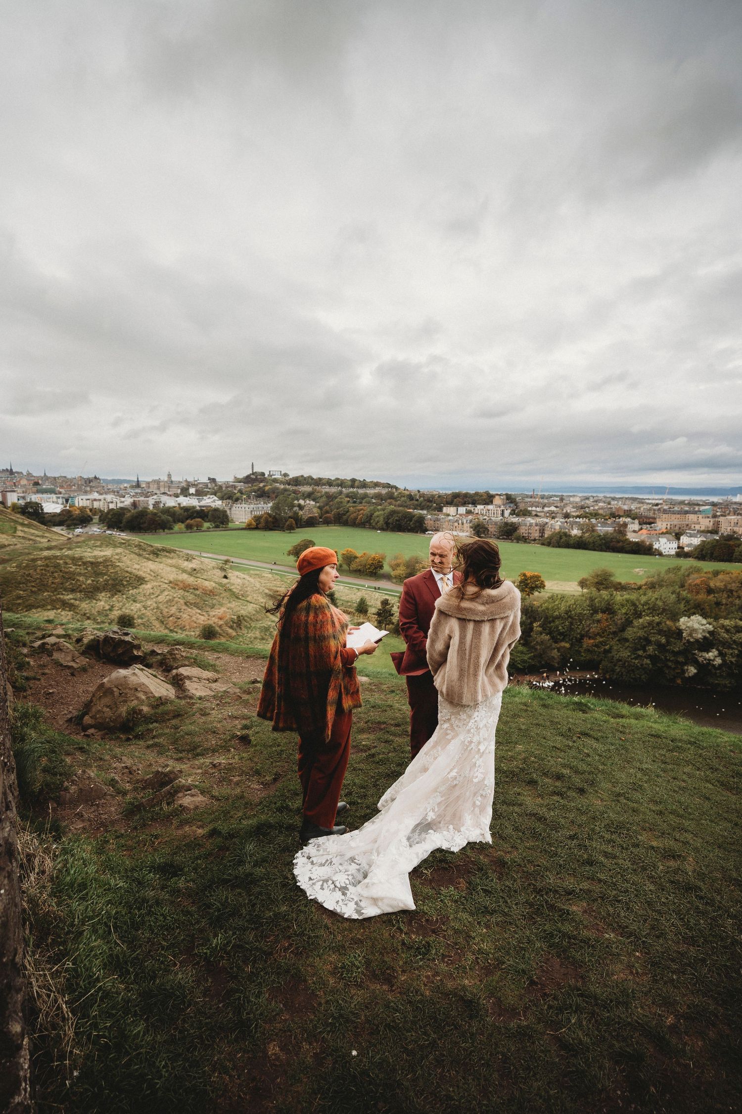 Wedding ceremony taking place on hilltop overlooking Edinburgh cityscape under dramatic sky.