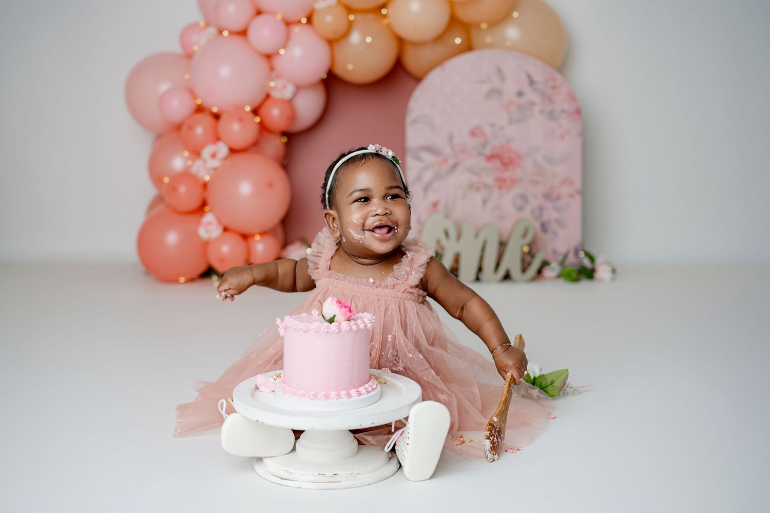 Baby girl in pink tutu dress smiles at first birthday cake smash with peach balloon arch backdrop.