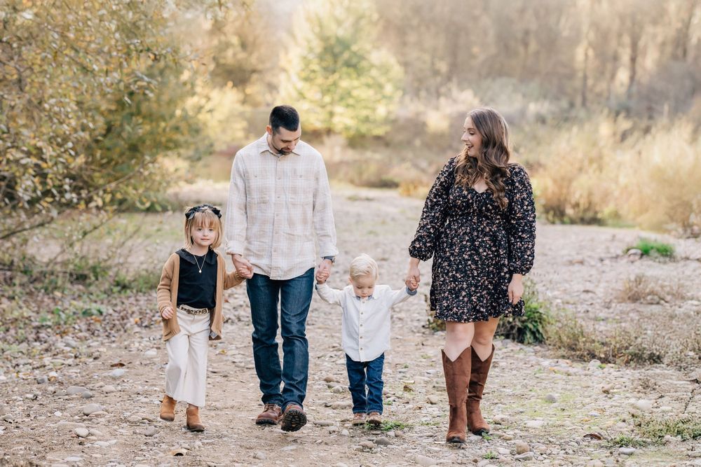 Family of four holding hands walking on a dirt path surrounded by autumn foliage during an outdoor photo session.