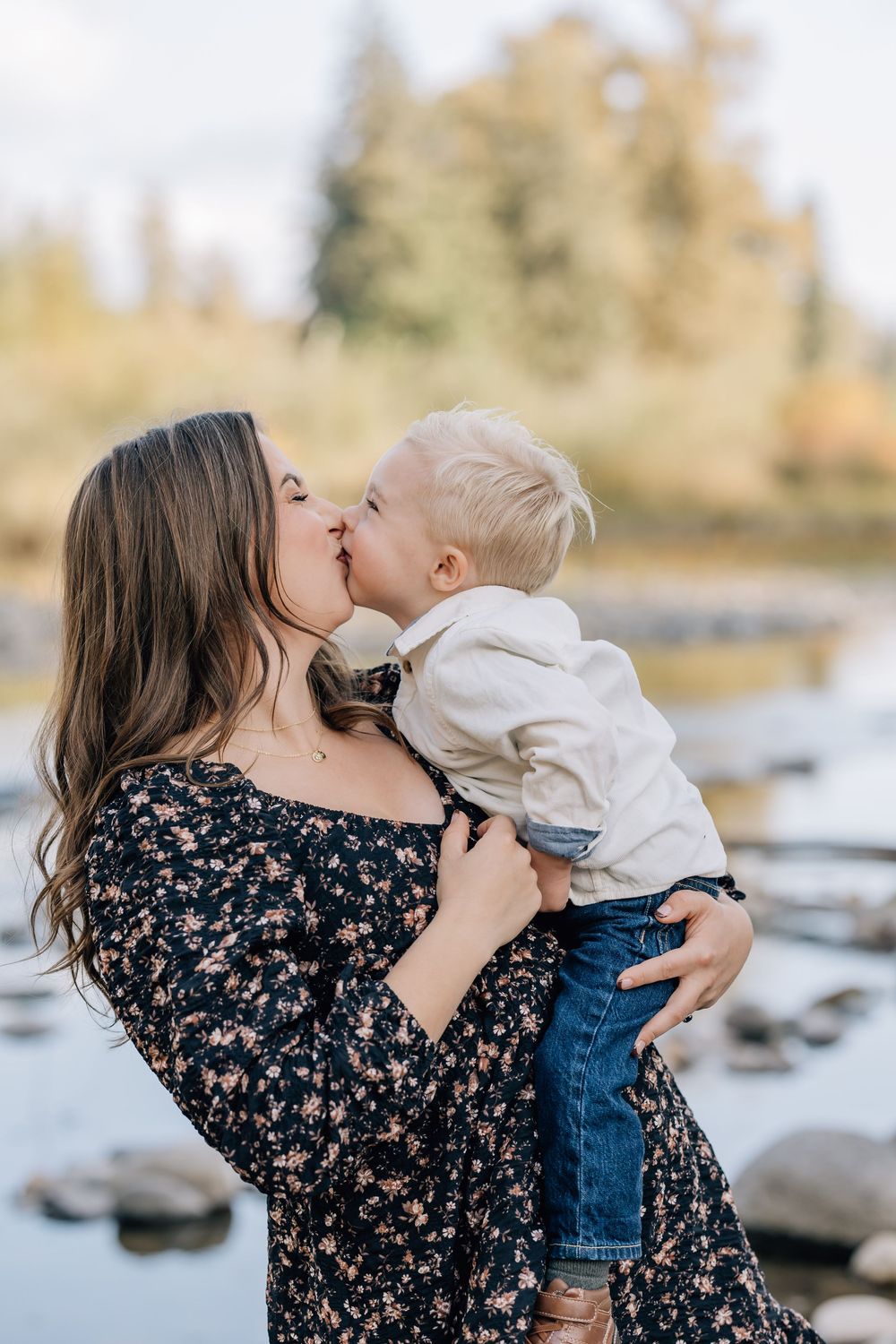 A mother holding and kissing her young blonde toddler son outdoors near a river in autumn.