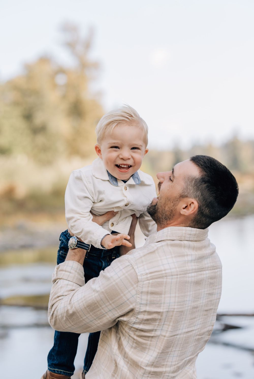 A smiling toddler being held by his father near a scenic river, both dressed in casual elegant outfits.