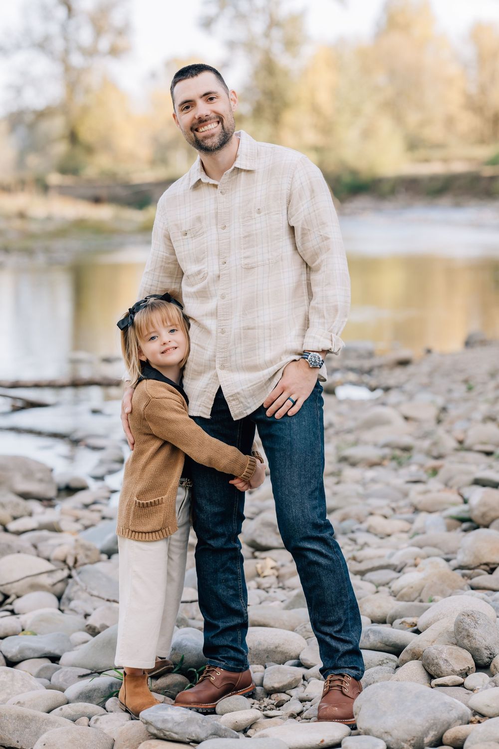 A smiling man and young girl pose together on a rocky riverbank surrounded by autumn trees.