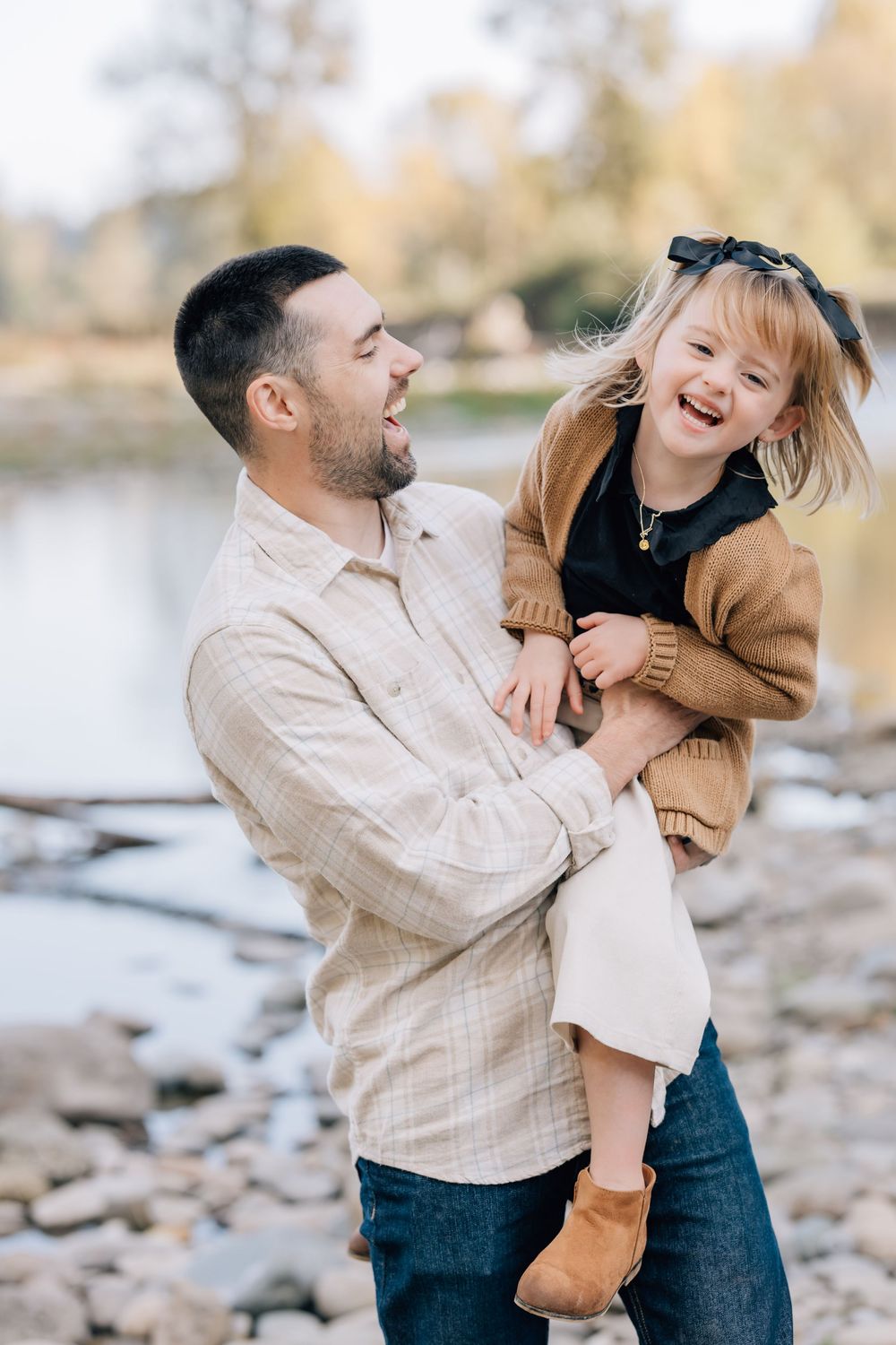 A laughing father holds his giggling young daughter outdoors near a rocky riverbank with autumn trees in the background.